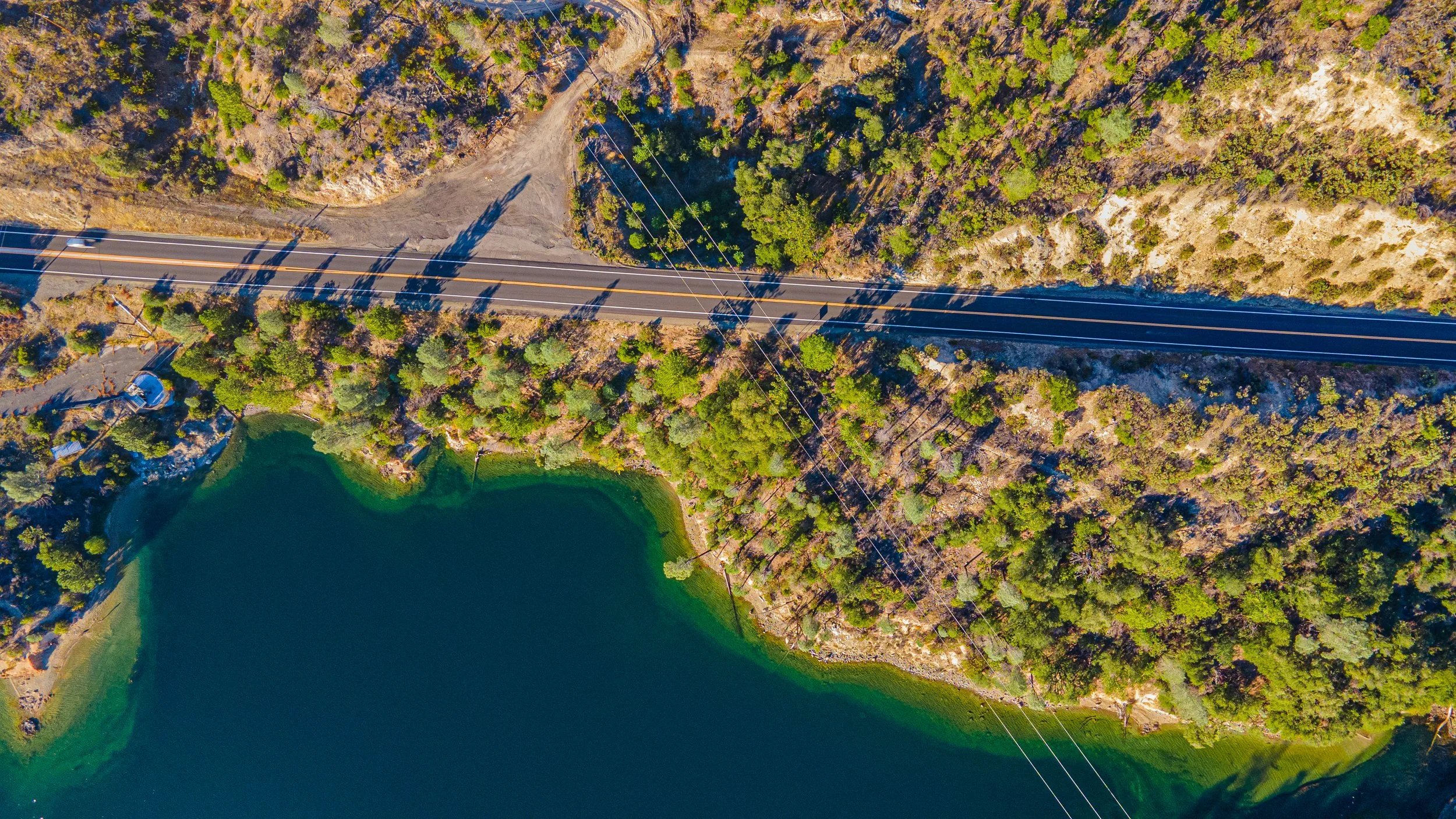 An aerial view of a winding road running alongside a body of water, surrounded by trees and rocky terrain.