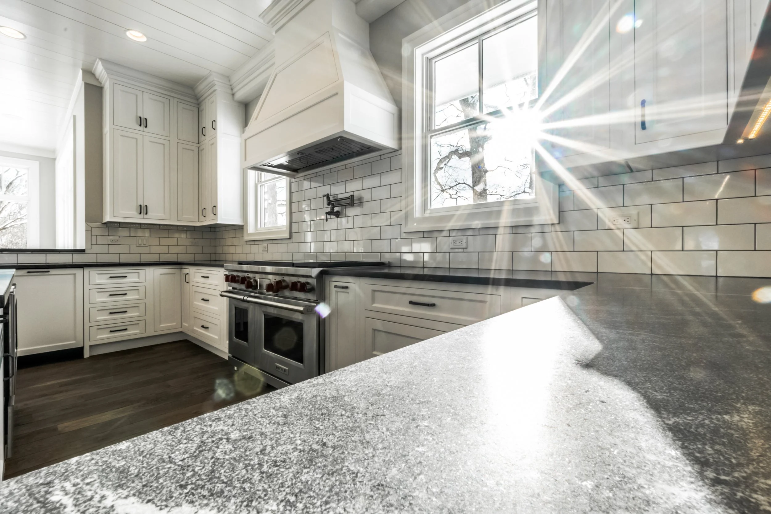 Bright kitchen with white cabinets, a black countertop, and a large window allowing sunlight to stream in, reflecting off a shiny backsplash of white subway tiles.
