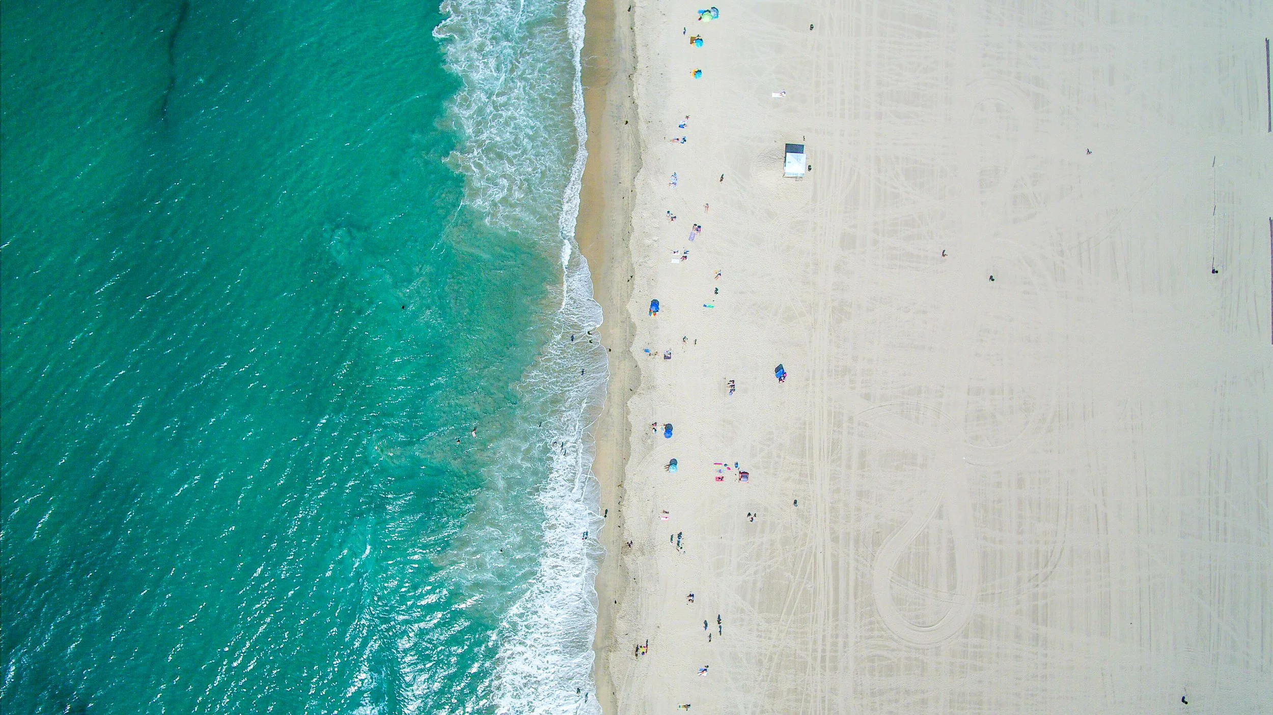 An aerial view of a beach with clear turquoise water, white sand, and people with umbrellas and chairs along the shore.