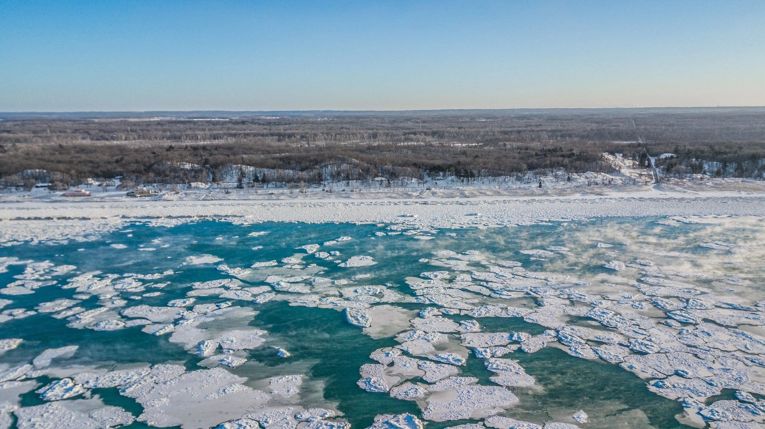 A frozen river with ice chunks floating on the surface, a snowy shoreline with trees, houses, and bare landscape under a clear blue sky.
