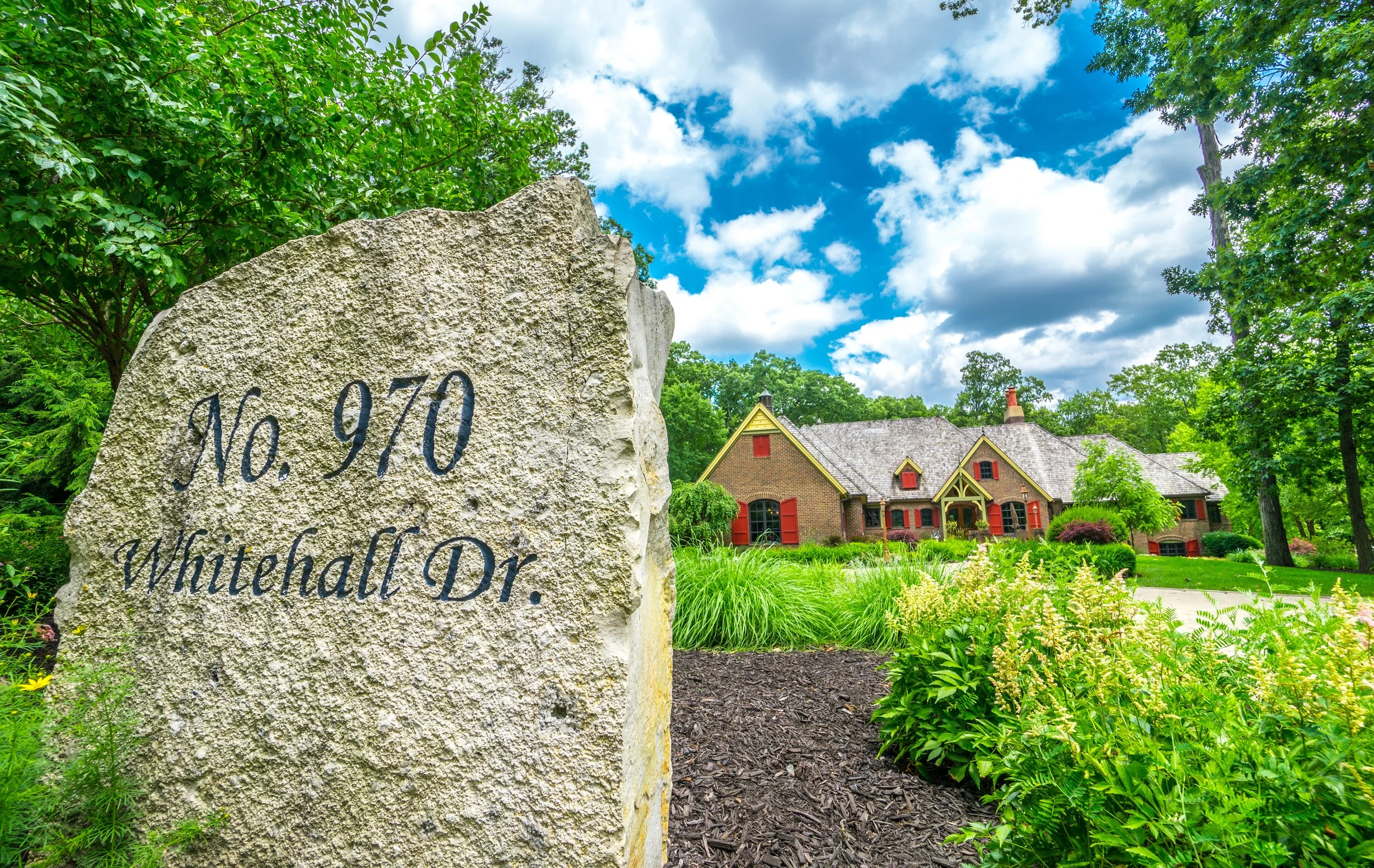 Stone marker with the text 'No. 970 Whitehall Dr.' in front of a house with brick exterior, red shutters, surrounded by lush green trees and landscaped garden, under a blue sky with scattered clouds.