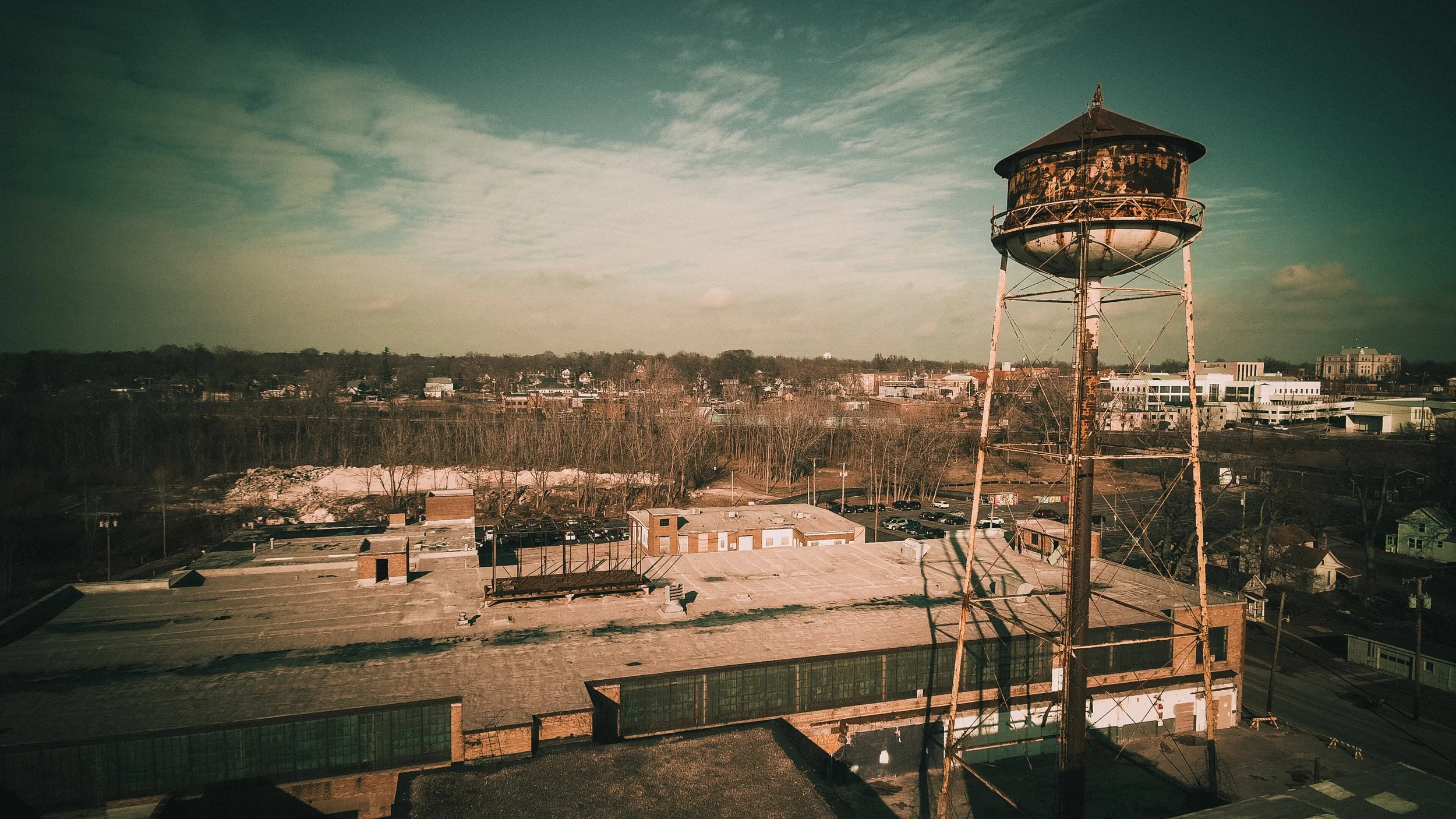 Old rusted water tower on a cityscape with industrial and residential buildings, parking lot, and a partly cloudy sky.