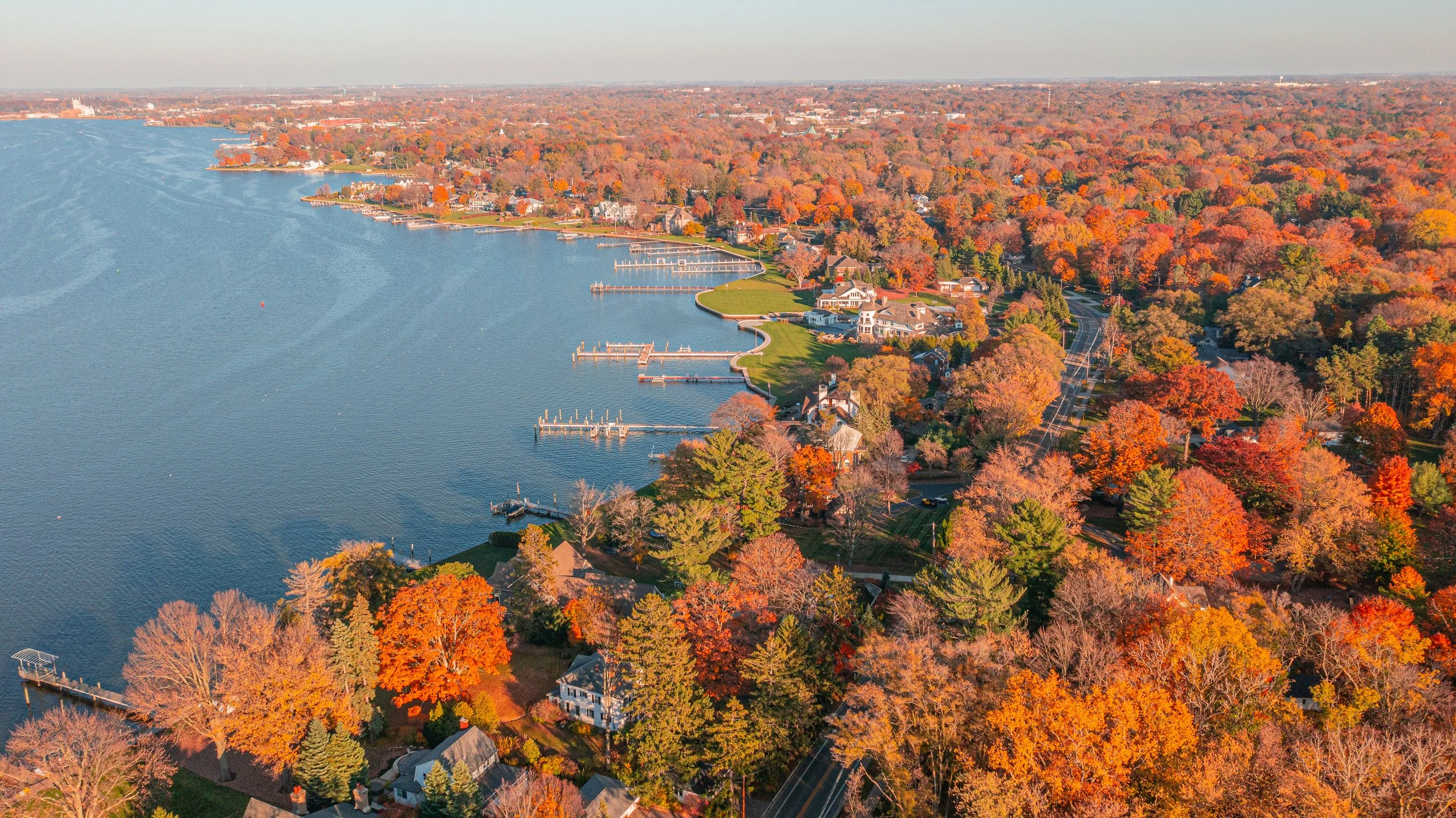 Aerial view of a lakeside neighborhood during autumn, showing colorful fall foliage, houses, docks, and winding streets.