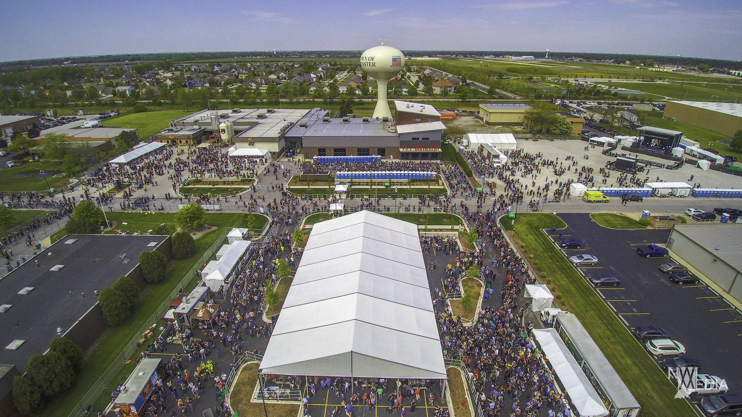 An aerial view of a large crowd gathered at an outdoor event near a stage, with tents, buildings, and a water tower in the background.