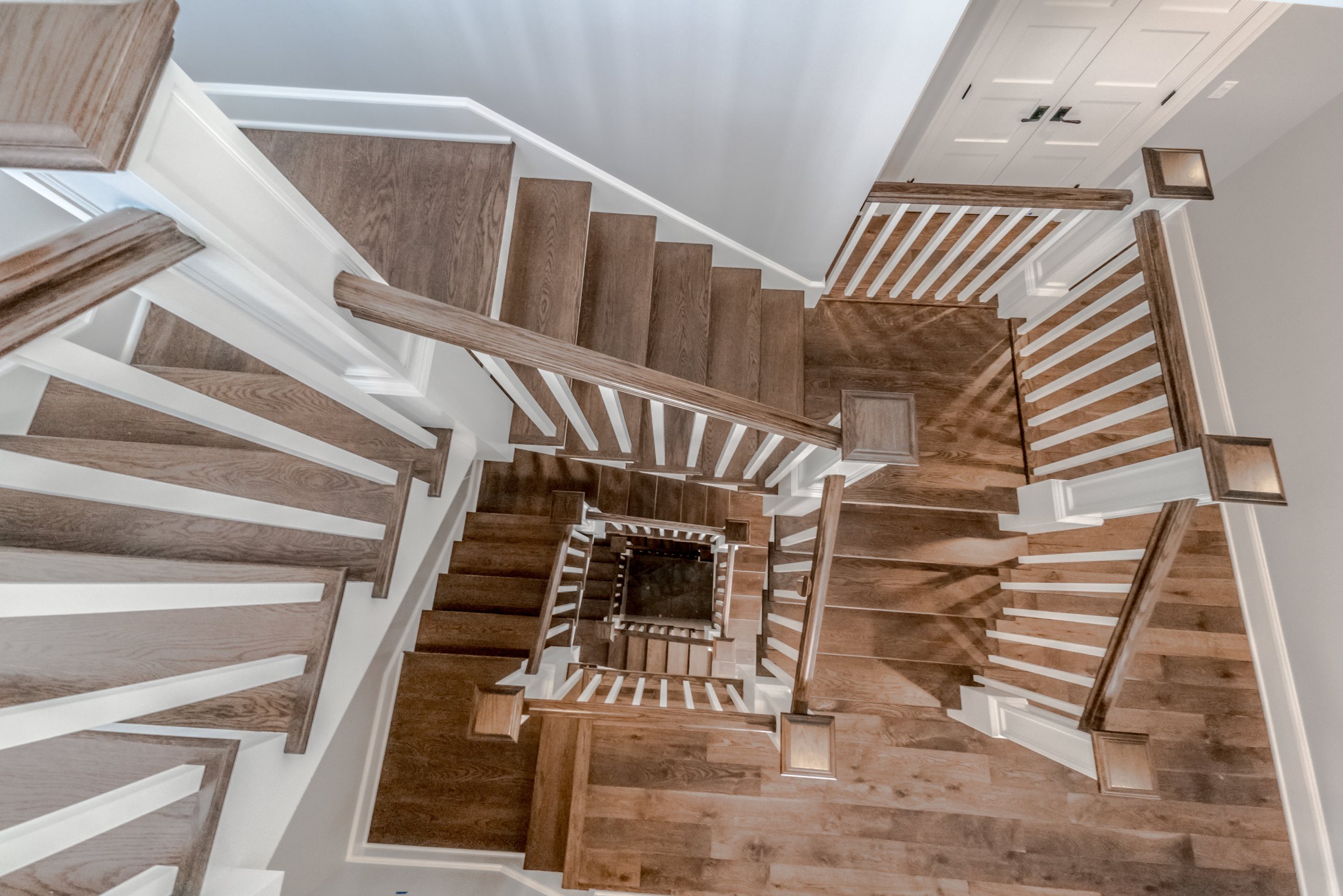 View down a staircase with brown wooden steps and white risers, white walls, and wooden handrails with white balusters.