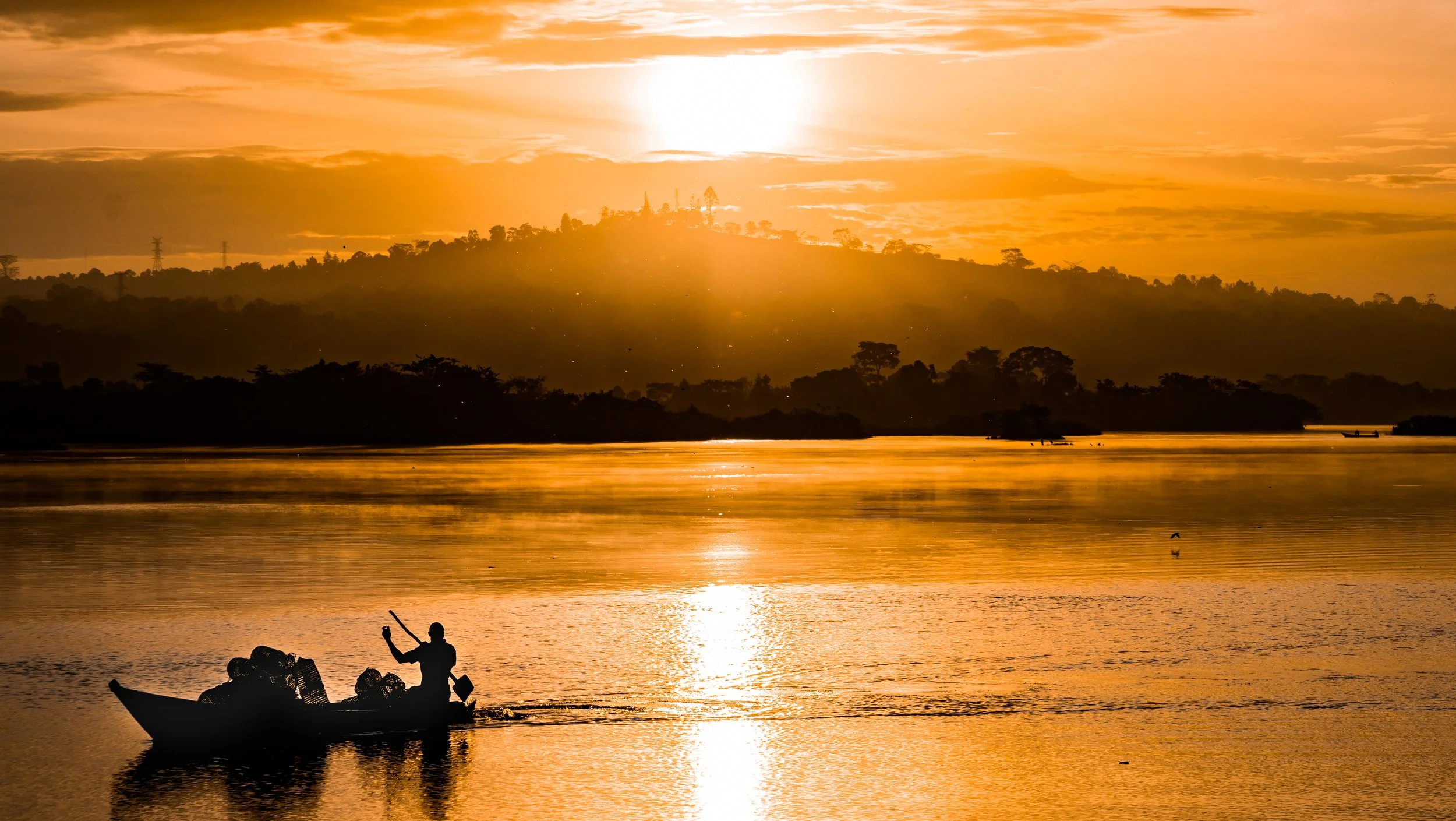 A person in a boat on a calm river during sunset with orange and yellow sky, silhouetted trees, and a person taking a photo or pointing at the sunset.