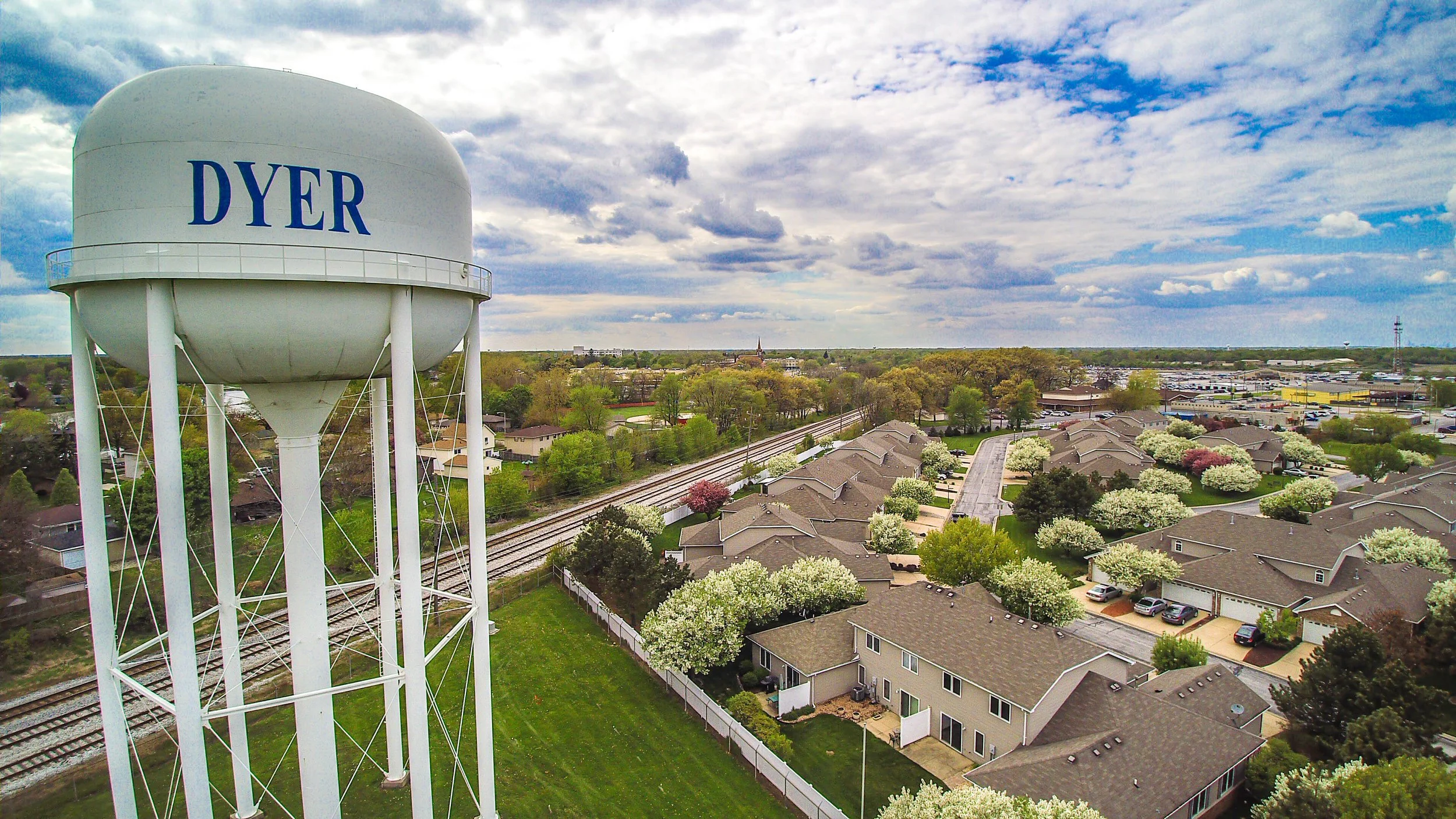 A water tower with the name 'DYER' on it stands above a residential neighborhood with houses, trees, and railway tracks in the background, under a partly cloudy sky.