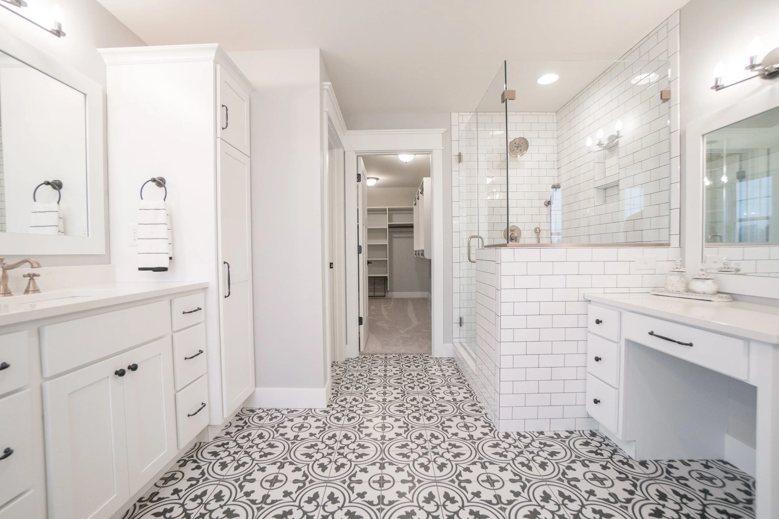 A bright, modern bathroom featuring white cabinetry, patterned black and white floor tiles, and a walk-in shower with white subway tile walls and a glass partition.