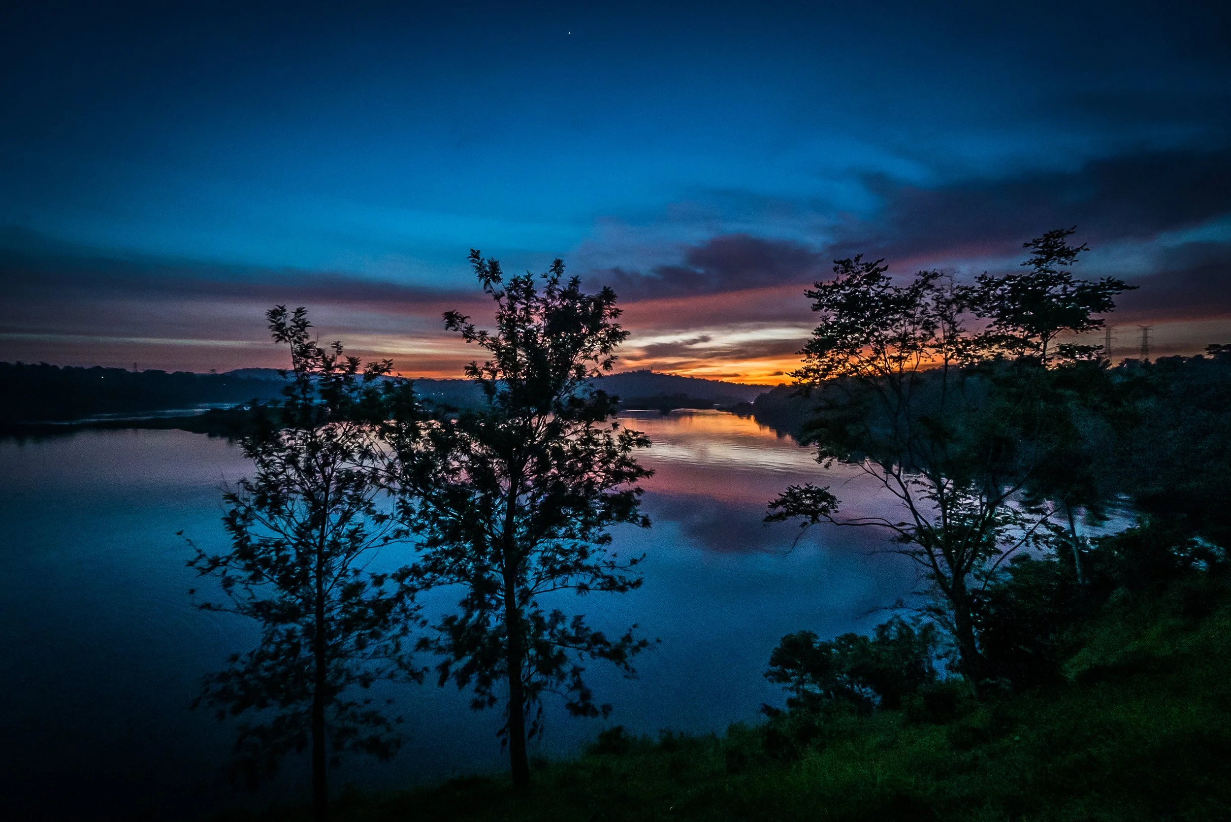 A river at dusk with a colorful sky showing shades of blue, pink, and orange, and trees silhouetted along the shoreline.