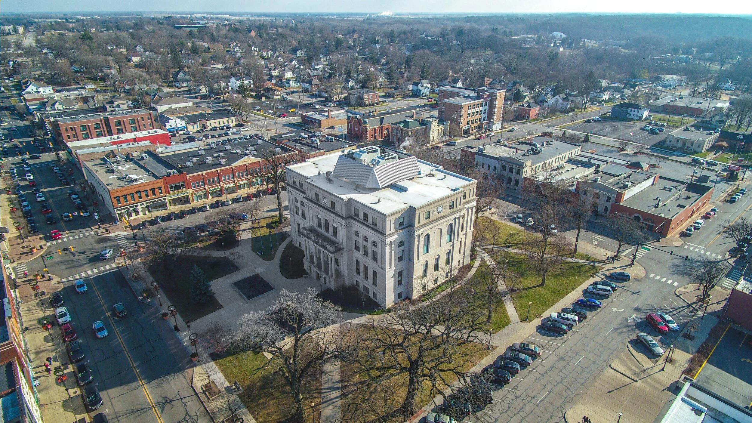 Aerial view of a downtown area with a prominent white classical-style building surrounded by parking lots, streets, and a mix of commercial and residential buildings, with leafless trees and some green patches.