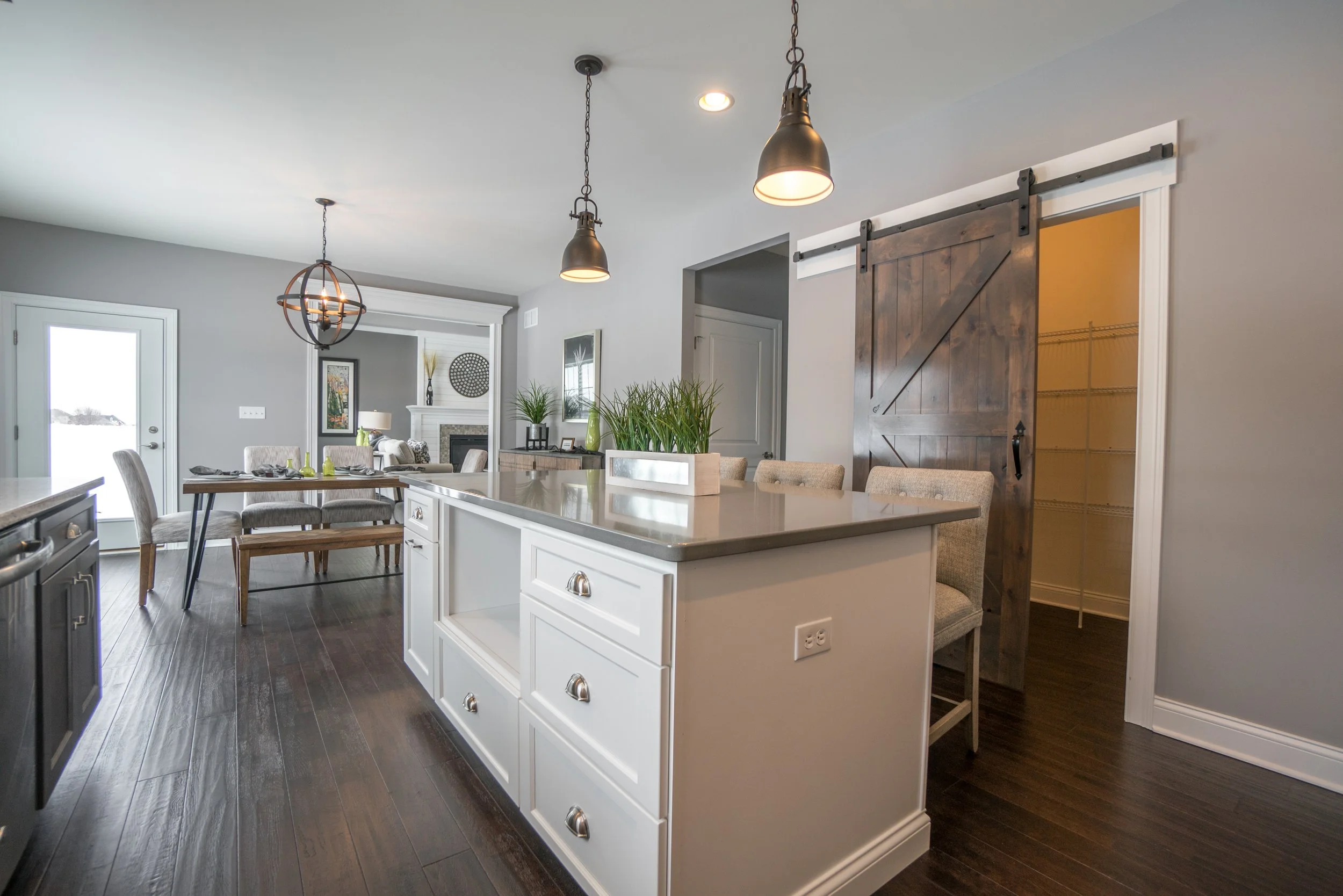 Modern kitchen and dining area with white cabinetry, a large island, wooden dining table with chairs, and a sliding barn door, illuminated by pendant lights.