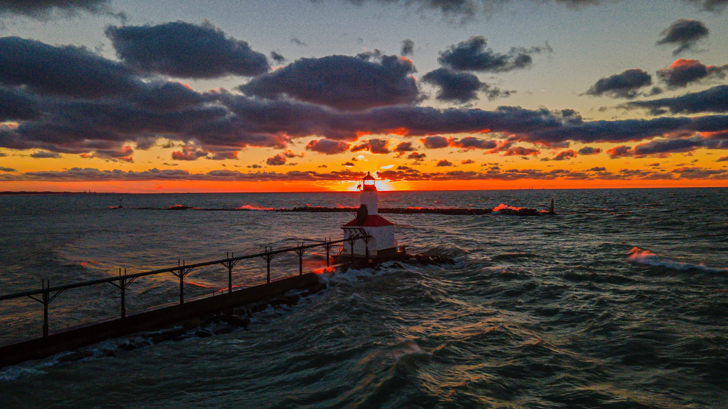 Sunset over the ocean with a lighthouse on a small rock jetty and a breakwater extending into the water.