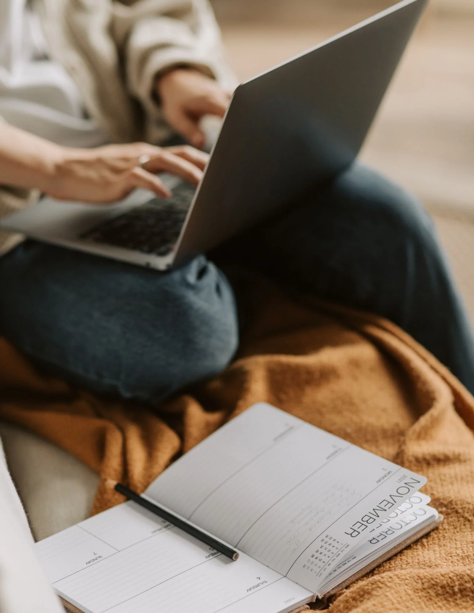 Person using a laptop beside an open planner and pen while sitting on a couch.