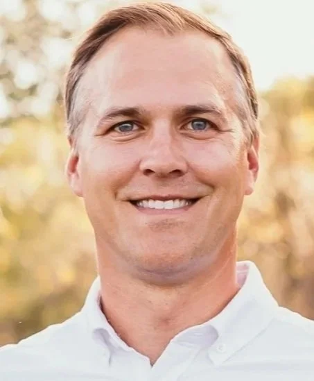 A smiling man outdoors with trees in the background, wearing a white shirt.