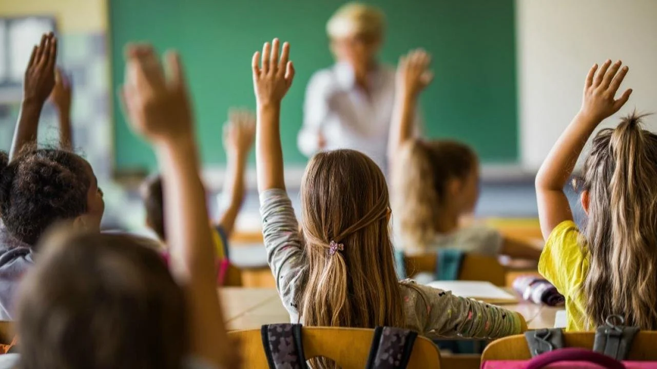 Students in a classroom raising their hands while a teacher stands in front of a green chalkboard.