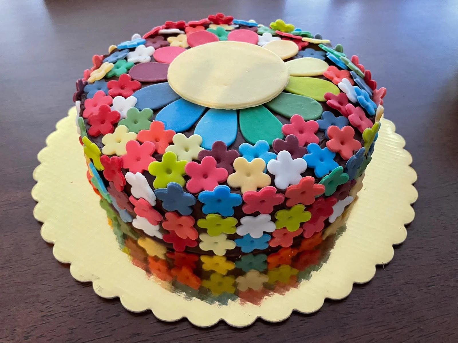 Colorful floral-shaped decoration on a round yellow cake, placed on a gold scalloped cake board on a dark wooden surface.