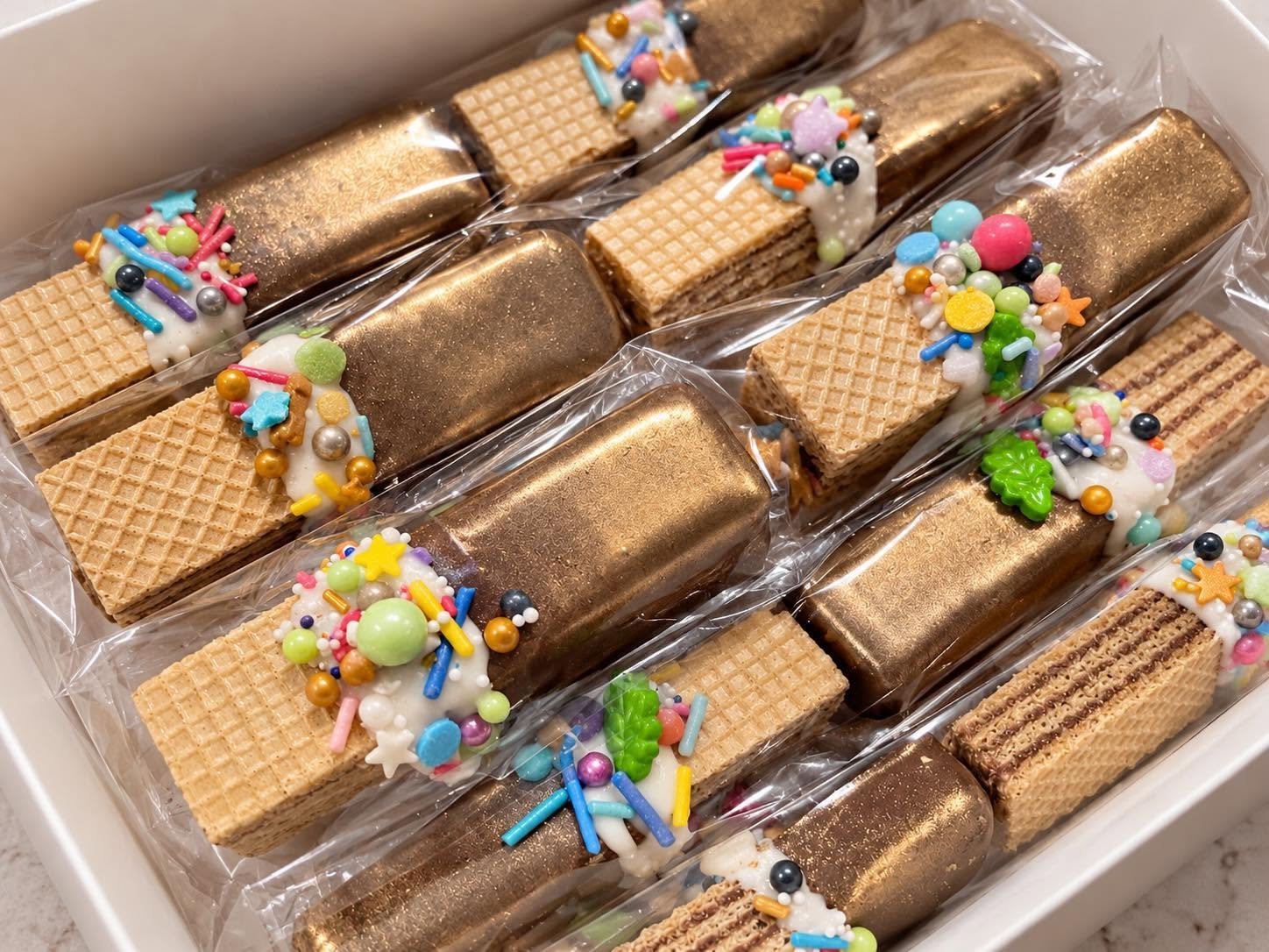 A box of wafer cookies decorated with colorful sprinkles and white icing, individually wrapped in clear plastic.
