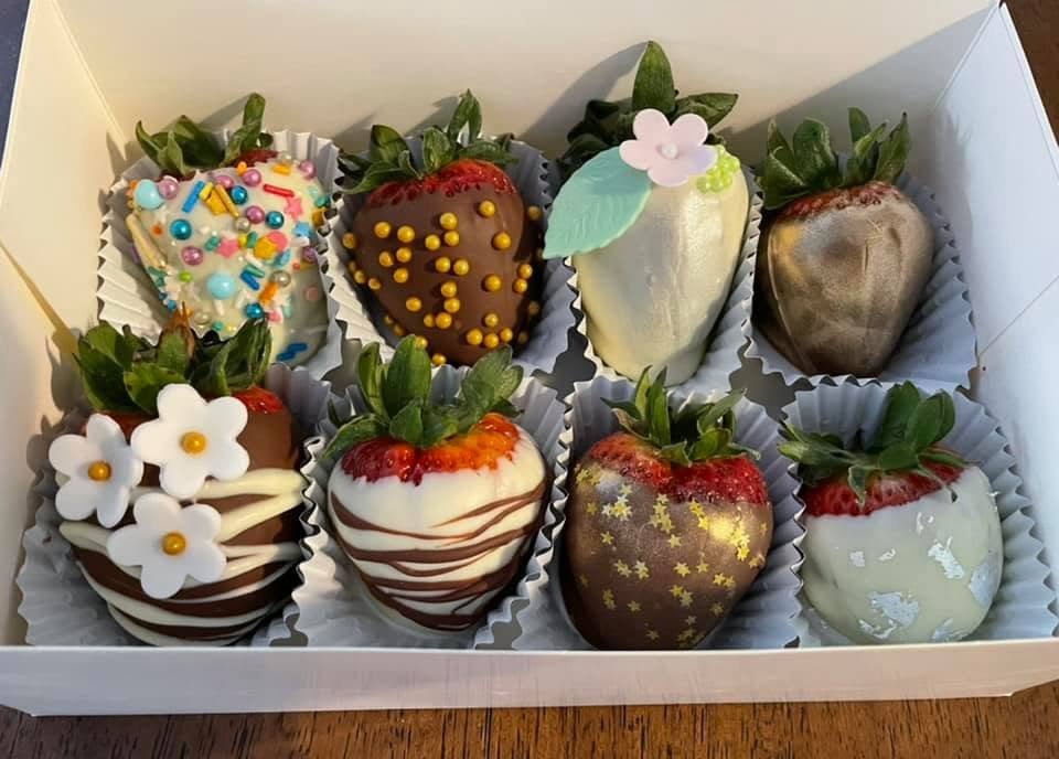 A box of eight decorated strawberries, some covered in chocolate, with various toppings including sprinkles, flowers, and white chocolate drizzle.