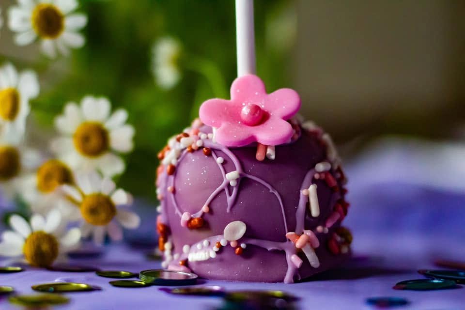 A purple cake ball decorated with colorful sprinkles and topped with a pink flower decoration and a white candle.