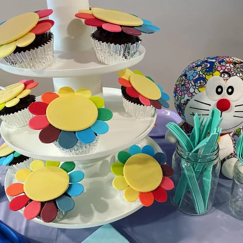 Colorful cupcake toppers resembling flowers, with a yellow center and petals in red, blue, green, brown, and pink, displayed on a white multi-tiered stand. A decorative plush toy of Doraemon, a Japanese robot cat, with a colorful patterned face and large eyes, sits beside a jar filled with teal-colored forks.