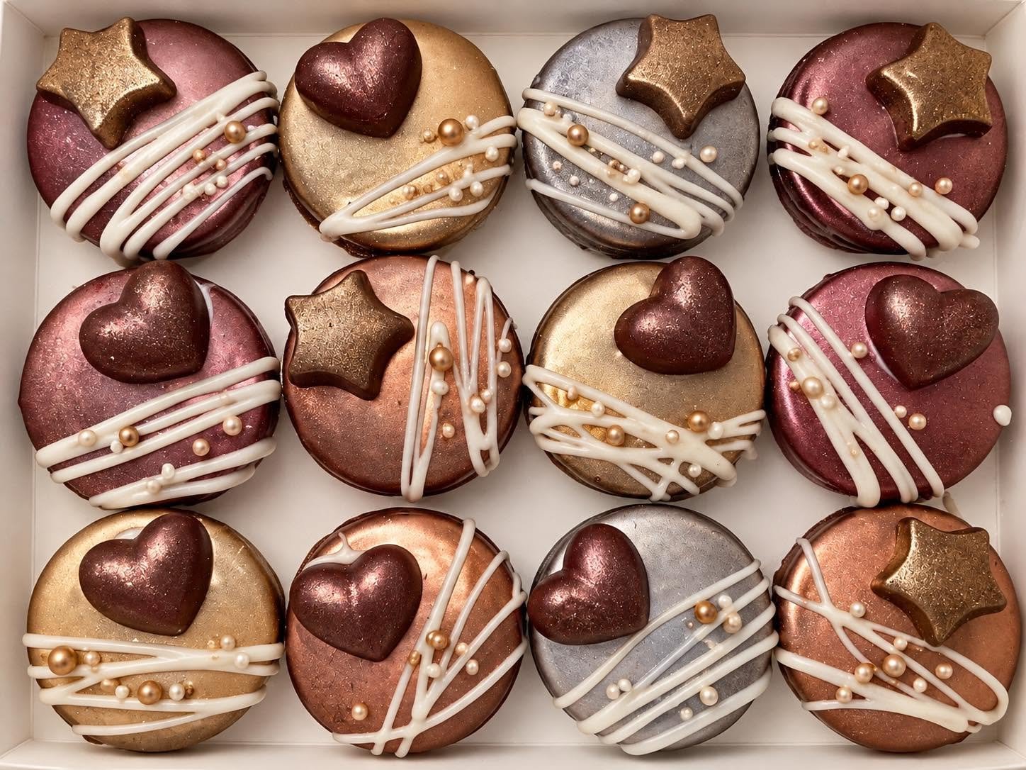 A box of 12 decorated cookies featuring metallic pink, gold, and silver colors, each topped with a heart or star-shaped decoration and white icing drizzles, gold and white edible pearls.