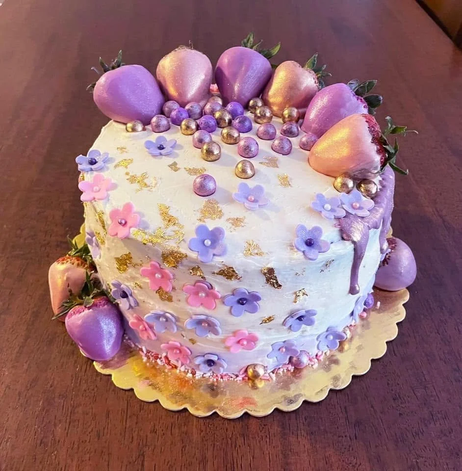 Decorated birthday cake with purple, pink, and gold strawberries, small purple and pink flowers, and gold accents on a gold cake board.