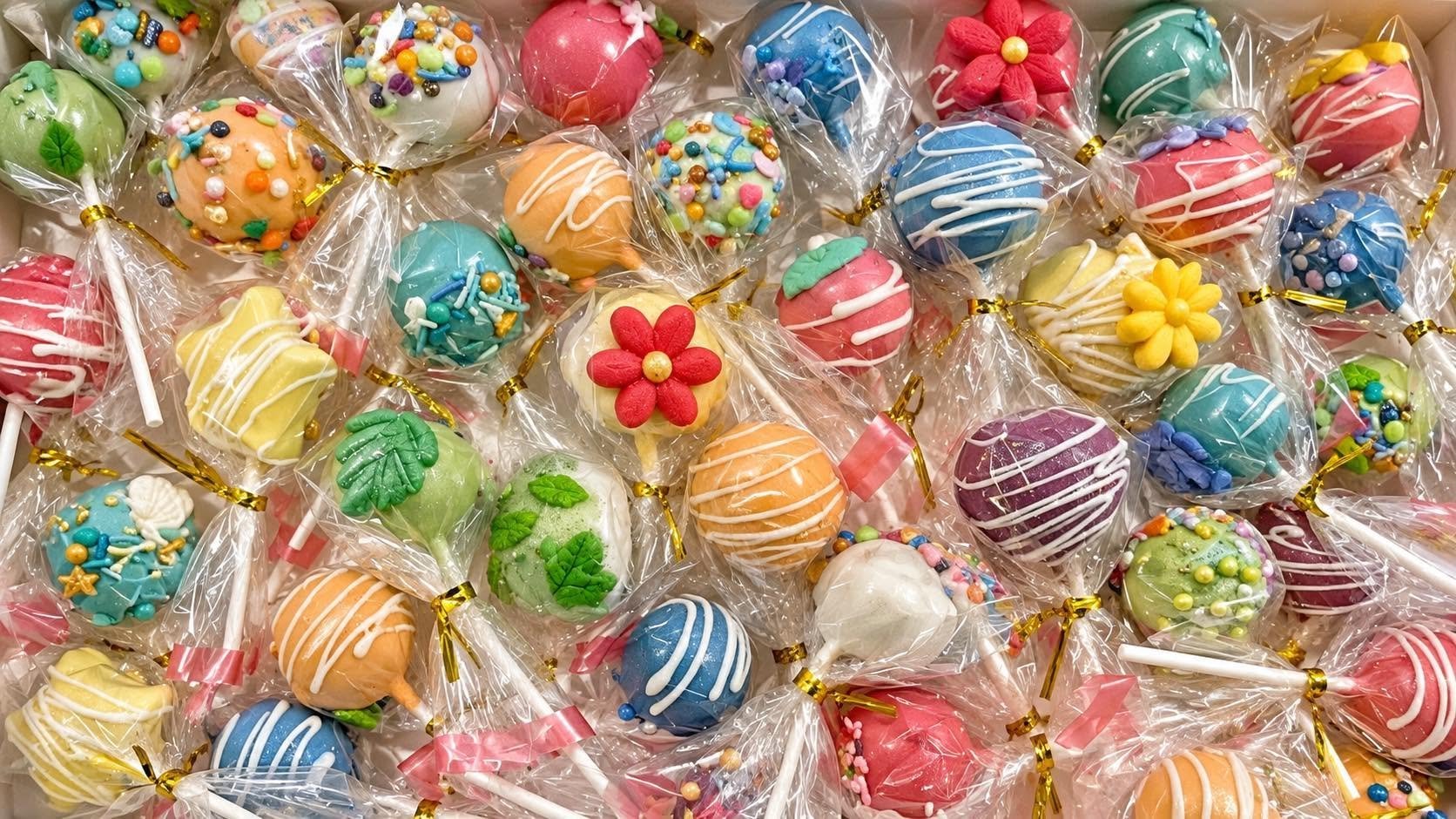 Colorful cake pops decorated with various sprinkles and icing, individually wrapped in clear plastic with gold twist ties.