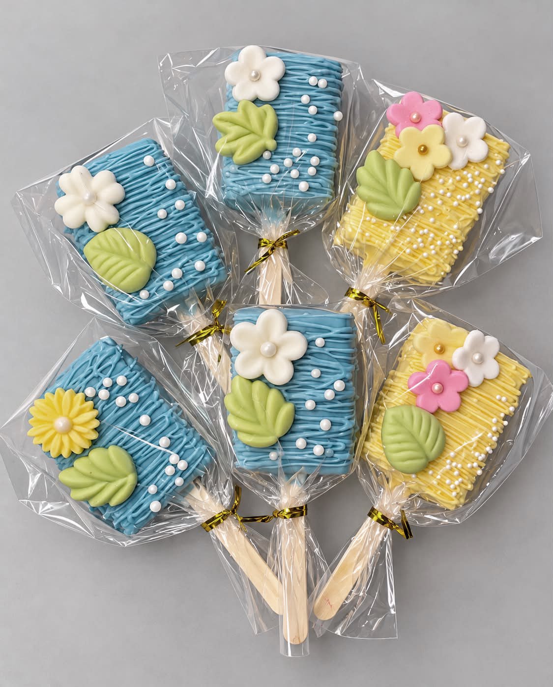 Colorful decorated cookies on sticks wrapped in clear plastic with gold twist ties, resembling popsicles with flowers and leaves, in blue and yellow icing