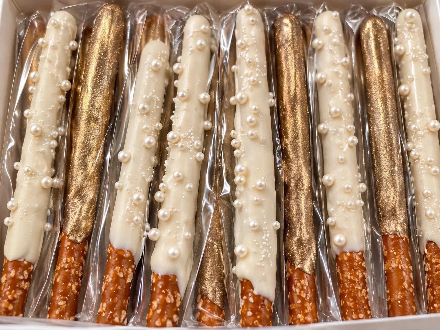 A box of decorated pretzel rods wrapped in clear plastic, with white and gold chocolate coatings, some have pearl-like sugar decorations and some have a metallic gold finish.