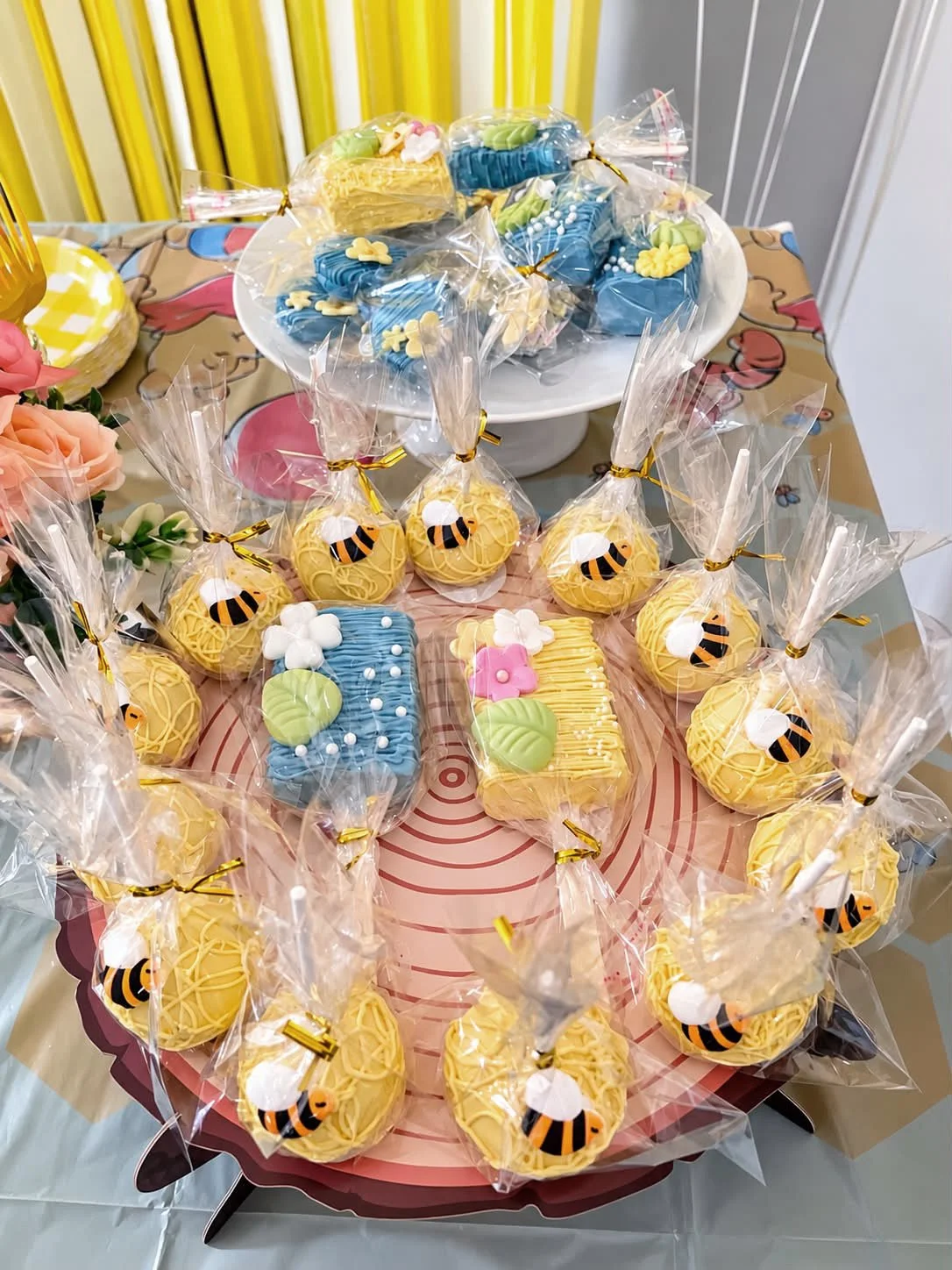 A table with yellow and blue decorated cookies, some wrapped in clear plastic bags tied with gold ribbons. The cookies are bee-themed with bee designs, and some are rectangular with pastel green leaves and pink flower accents.