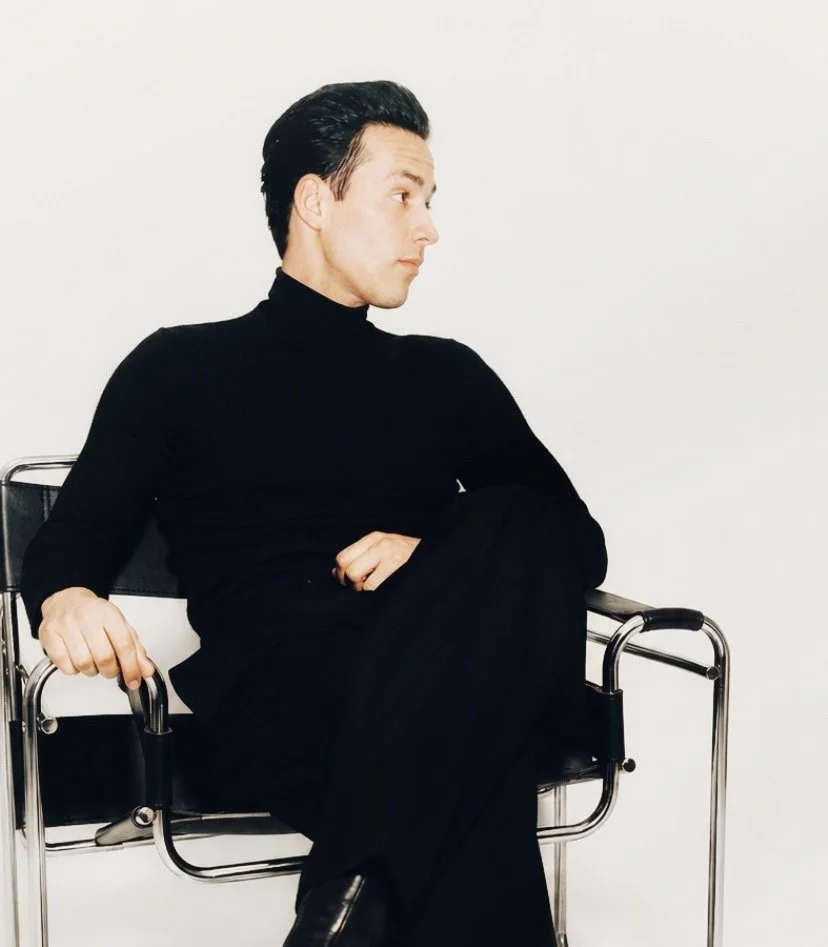 A young man with dark hair sitting on a black chair, wearing a black turtleneck and black pants, looking to the side with a neutral expression against a plain white background.
