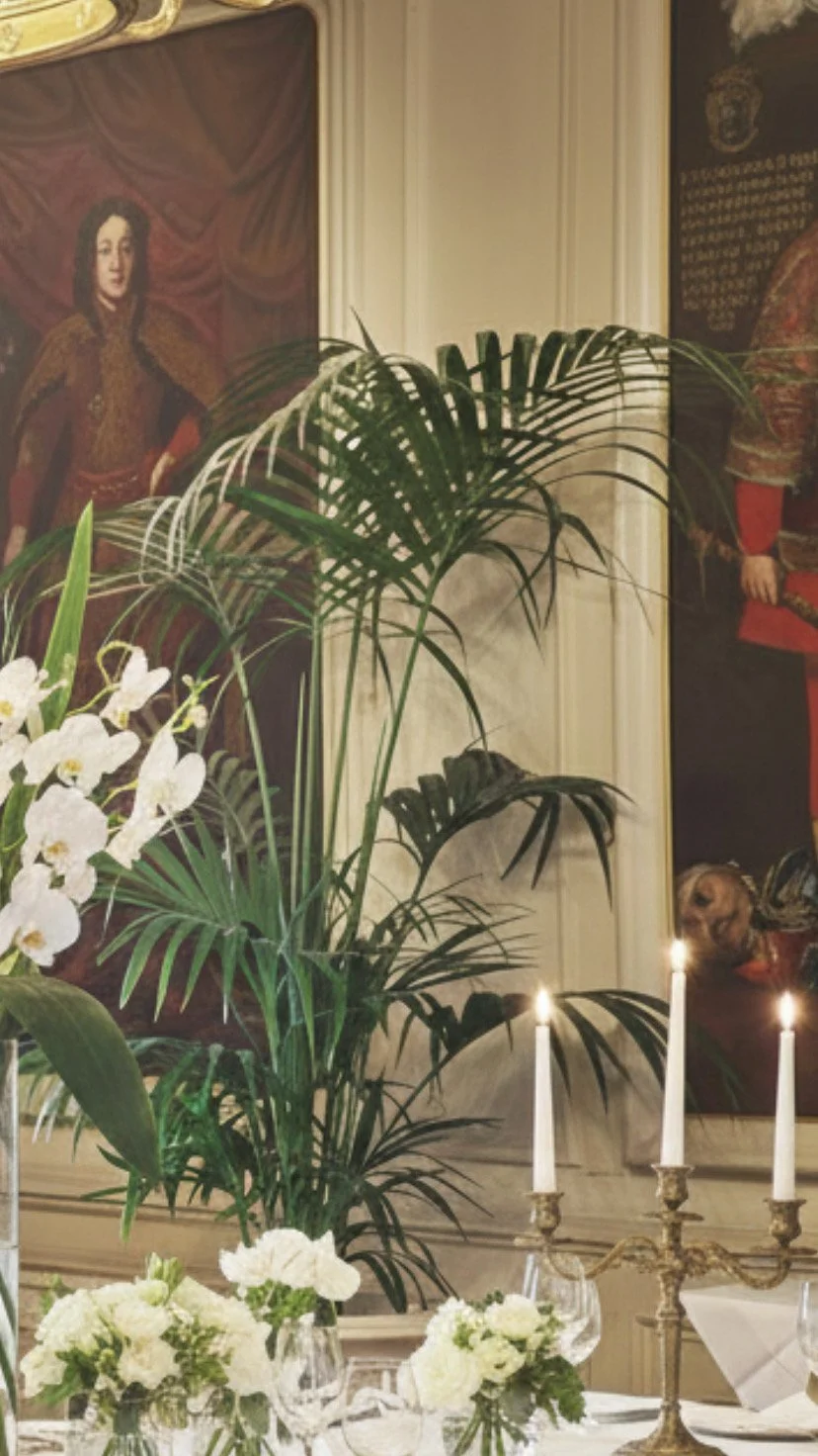 Elegant table setting with white floral arrangements, a candelabra with three lit candles, and large green leafy plants in the background, inside a room with historical paintings on the wall.