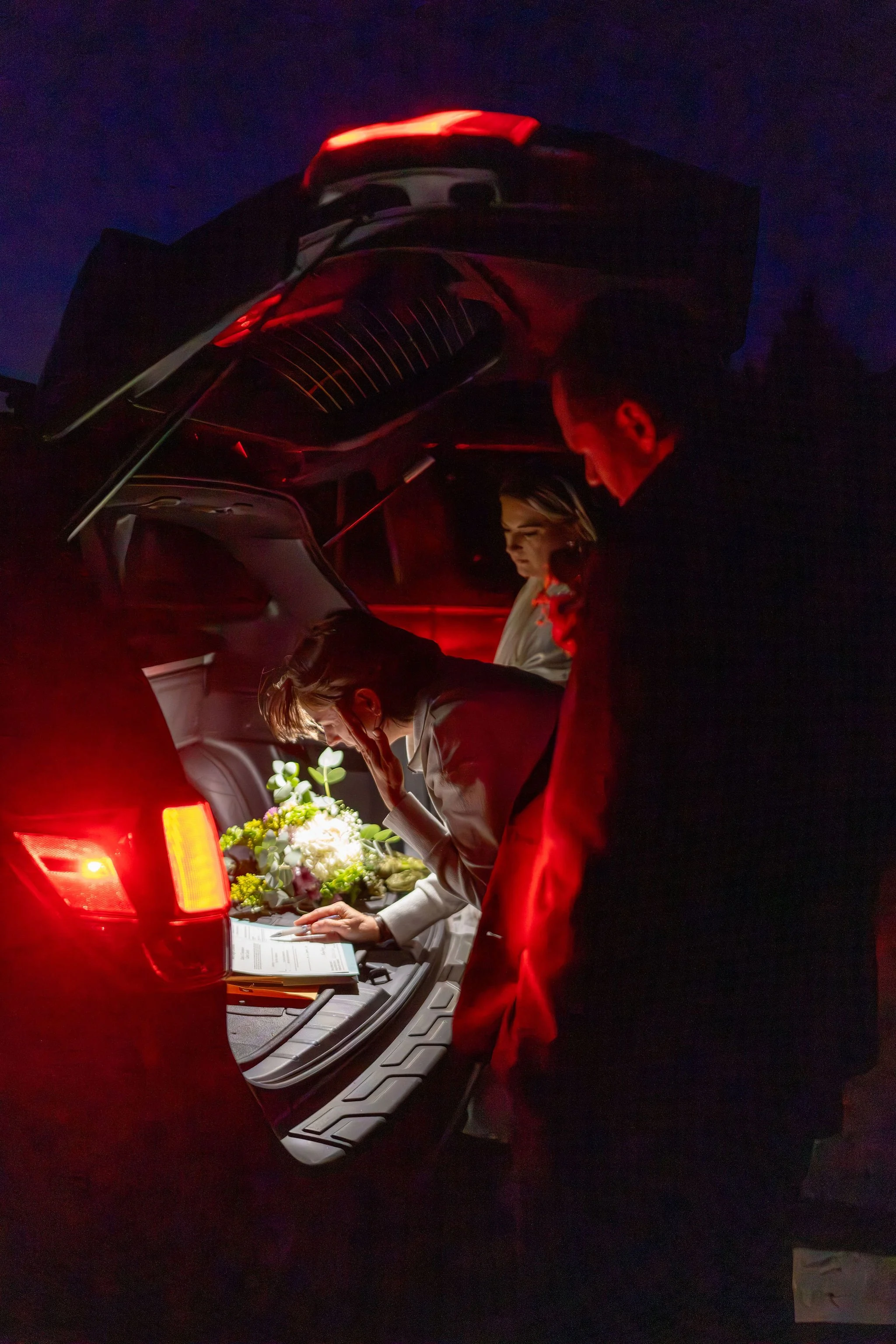 Officiating the wedding with the open trunk of a car at night, with one person placing flowers inside, illuminated by red and ambient lighting.
