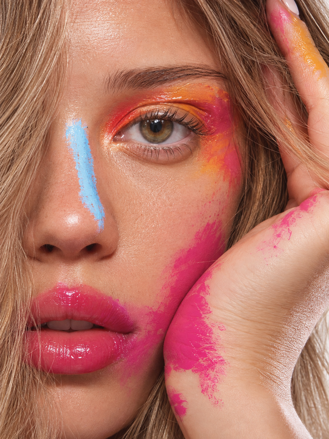 Close-up of a woman's face with colorful makeup and paint splashes, one eye visible, her hand resting near her face with pink paint on her fingers.
