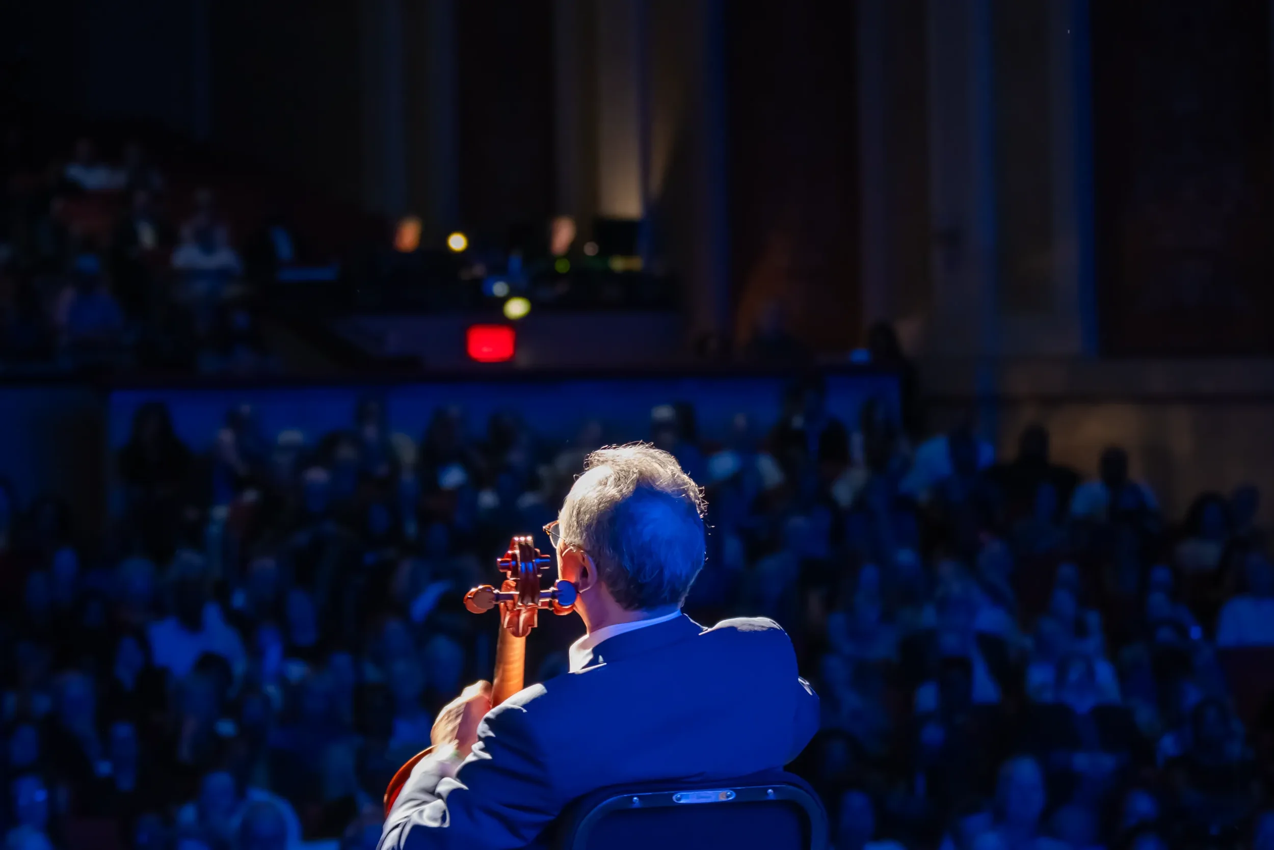 A person playing a violin on stage in front of a large audience, with blue stage lighting and curtains in the background.