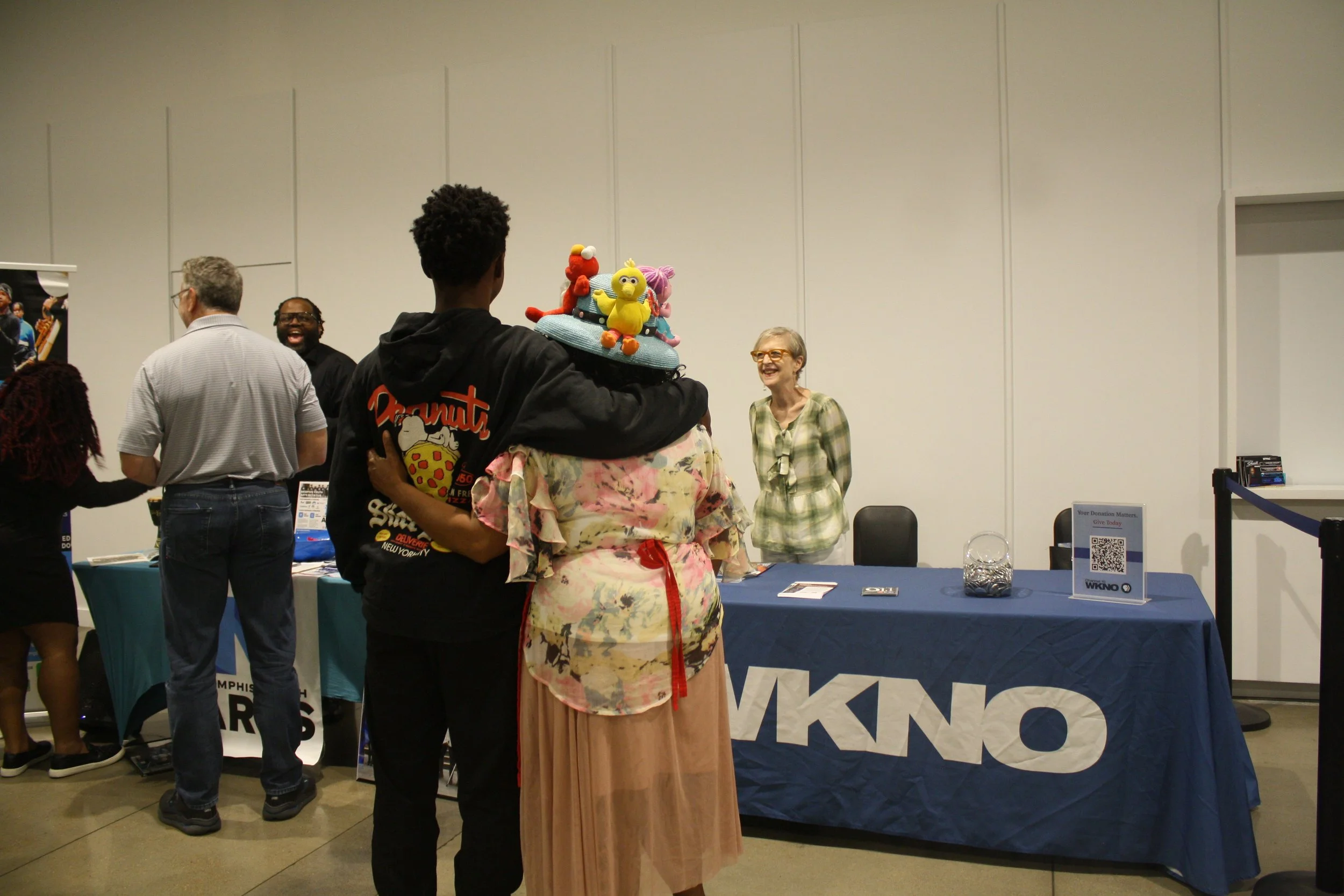 People at a booth or event, some engaging with each other, with a woman wearing a large colorful hat with puppet figures, and a woman smiling in a plaid shirt in the background.
