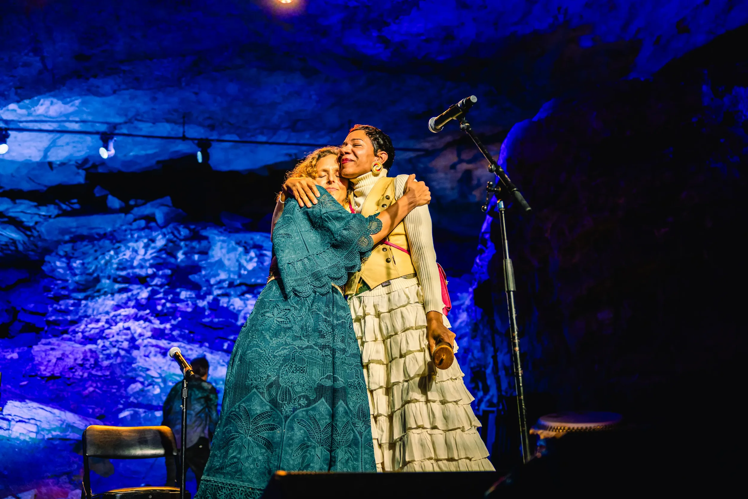 Two women embracing each other on stage during a performance, with a microphone stand nearby, in front of a rocky blue-lit background.