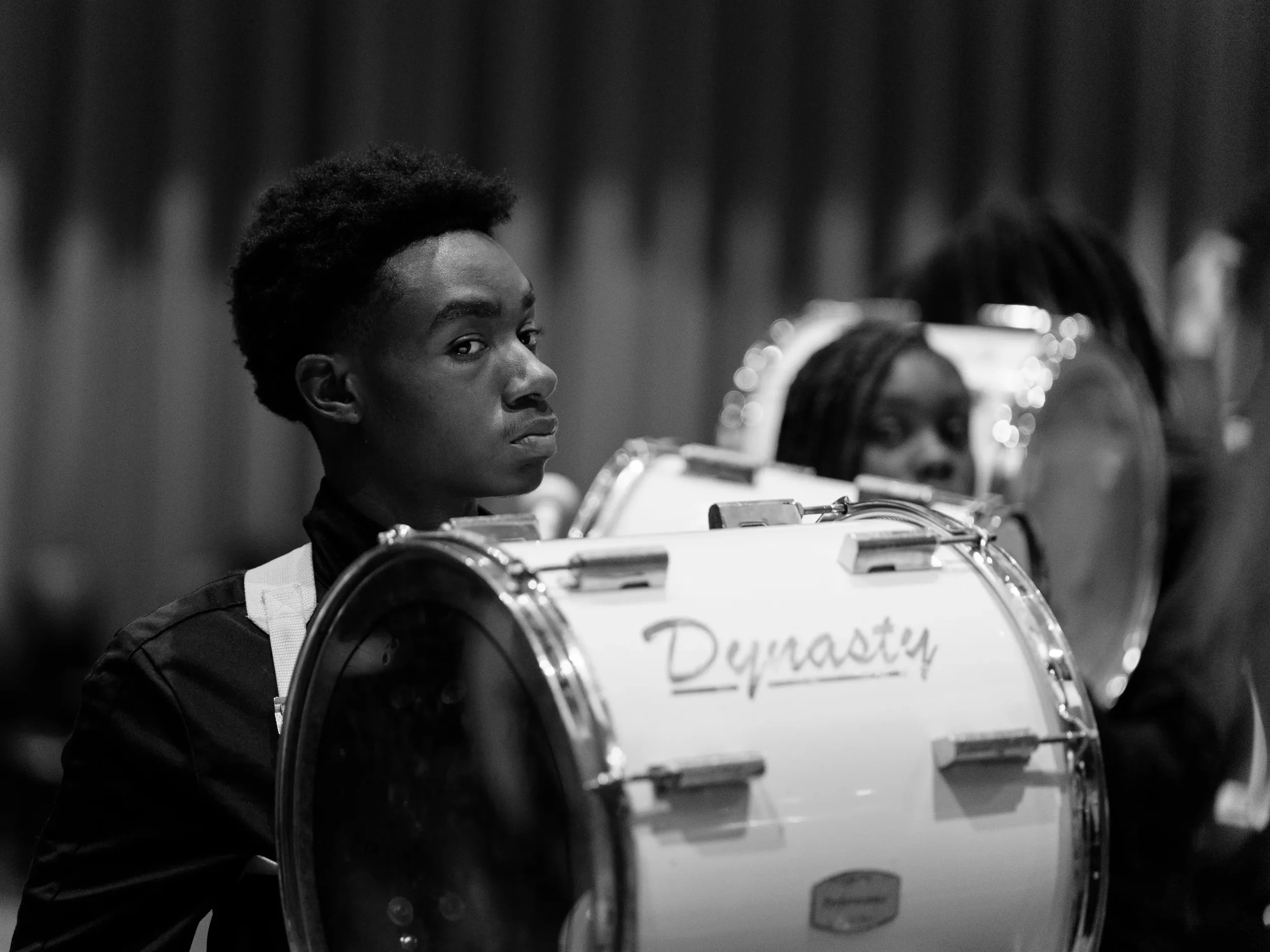 A young boy is sitting behind a drum with a sign that says 'Drama T.' There are other people, including a girl with glasses and her hair in dreadlocks, in the background.