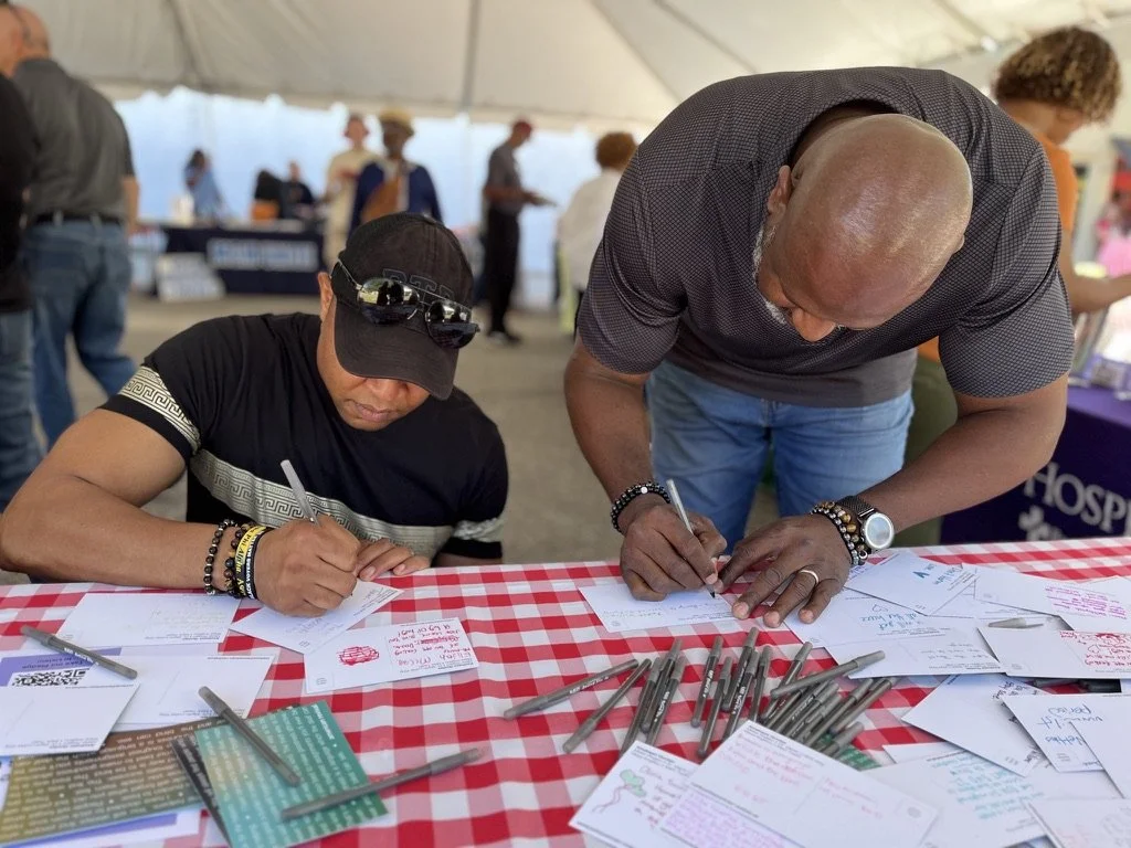 Two men signing papers on a red and white checkered table at an event with people in the background.