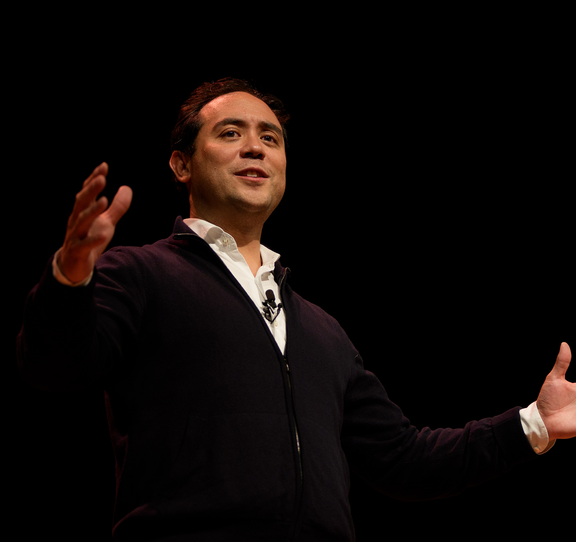 A man speaking on stage, gesturing with his right hand, wearing a dark zip-up sweater over a white shirt, set against a black background.