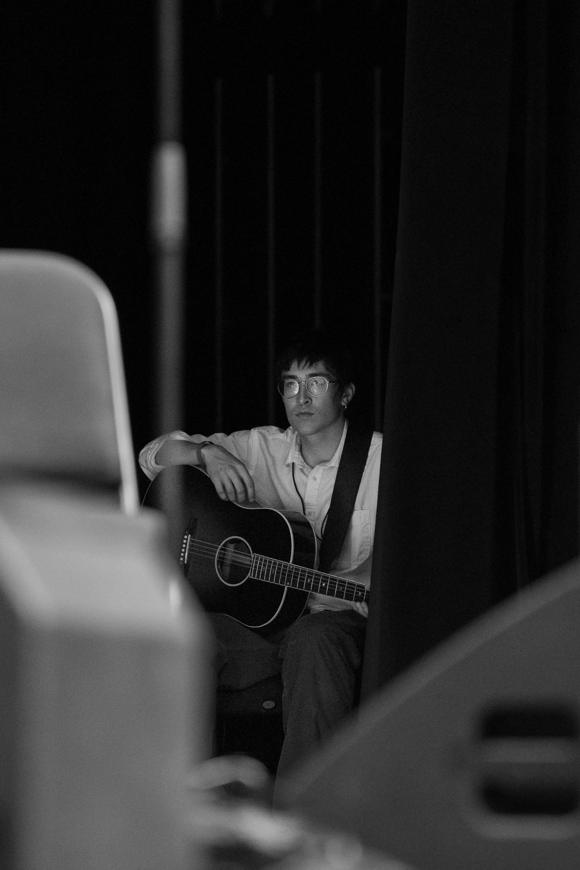 A person wearing glasses and a light-colored shirt, sitting in a dark room, playing an acoustic guitar, possibly performing or practicing.