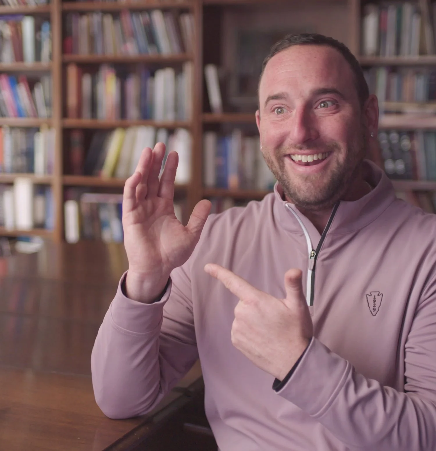A smiling man with a beard and short hair, wearing a light pink sports jacket, is sitting at a wooden table and gesturing with his hands. He is in a room with wooden bookshelves filled with books in the background.
