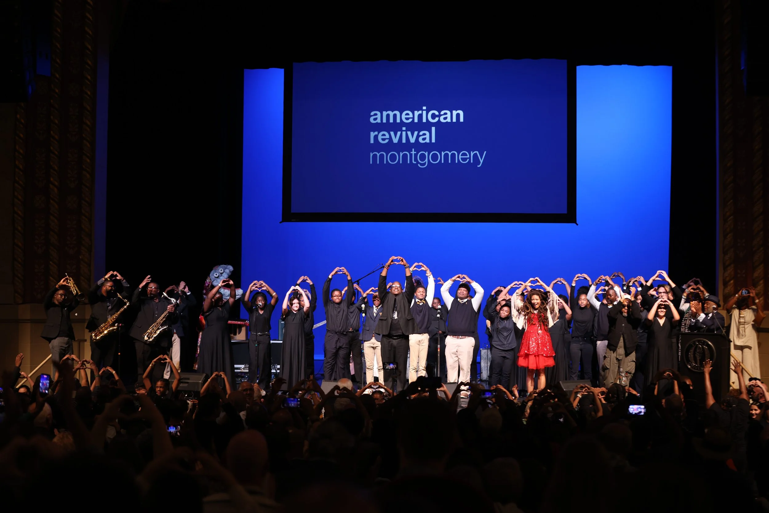 A group of children and young adults on stage at the American Revival event in Montgomery, some holding microphones and musical instruments, forming heart shapes with their hands, with a large screen displaying the event name behind them.