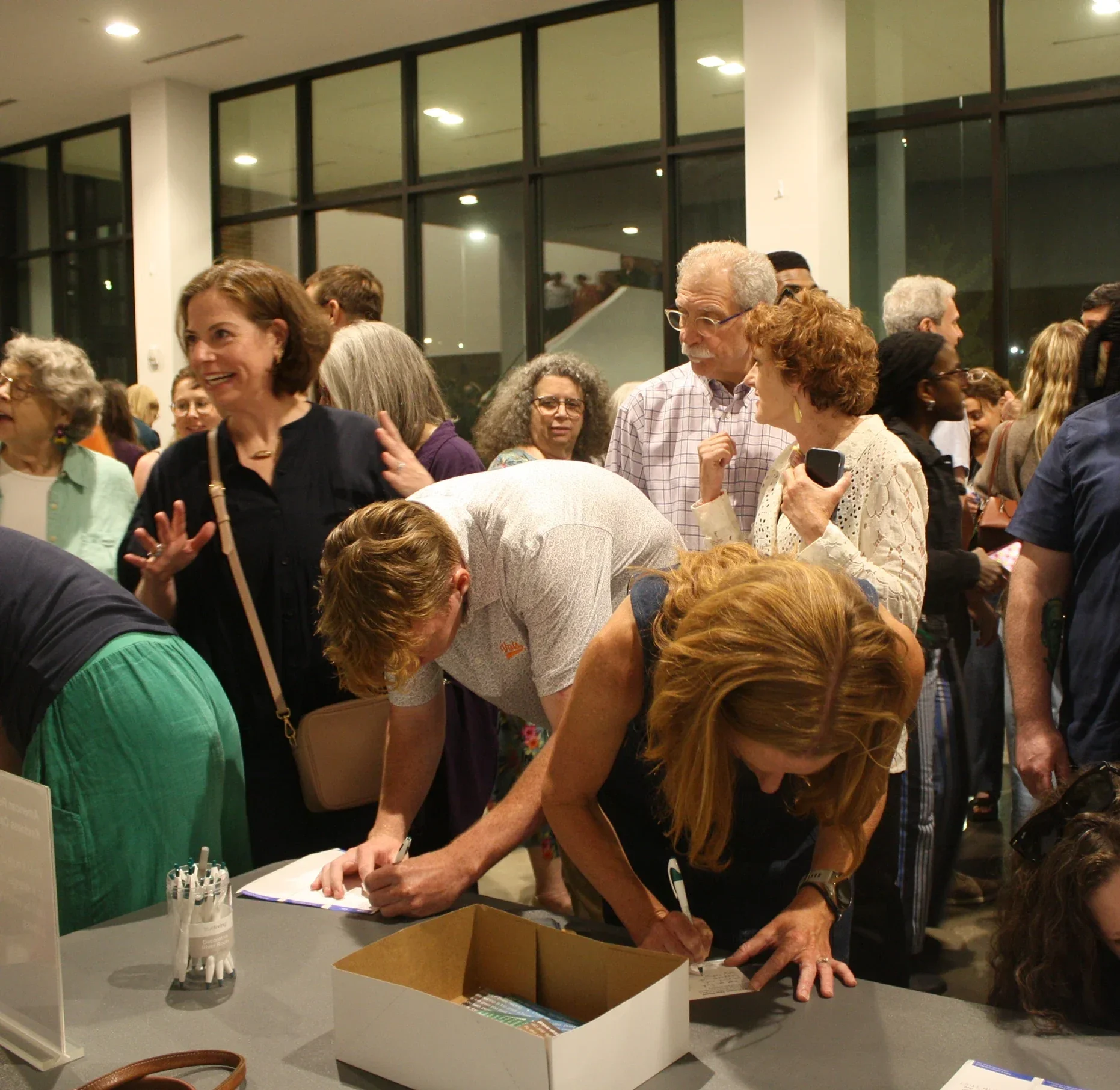 People gather around a table at an indoor event, signing papers with pens. The scene is busy, with individuals engaging and interacting. The background shows large glass windows and multiple overhead lights.