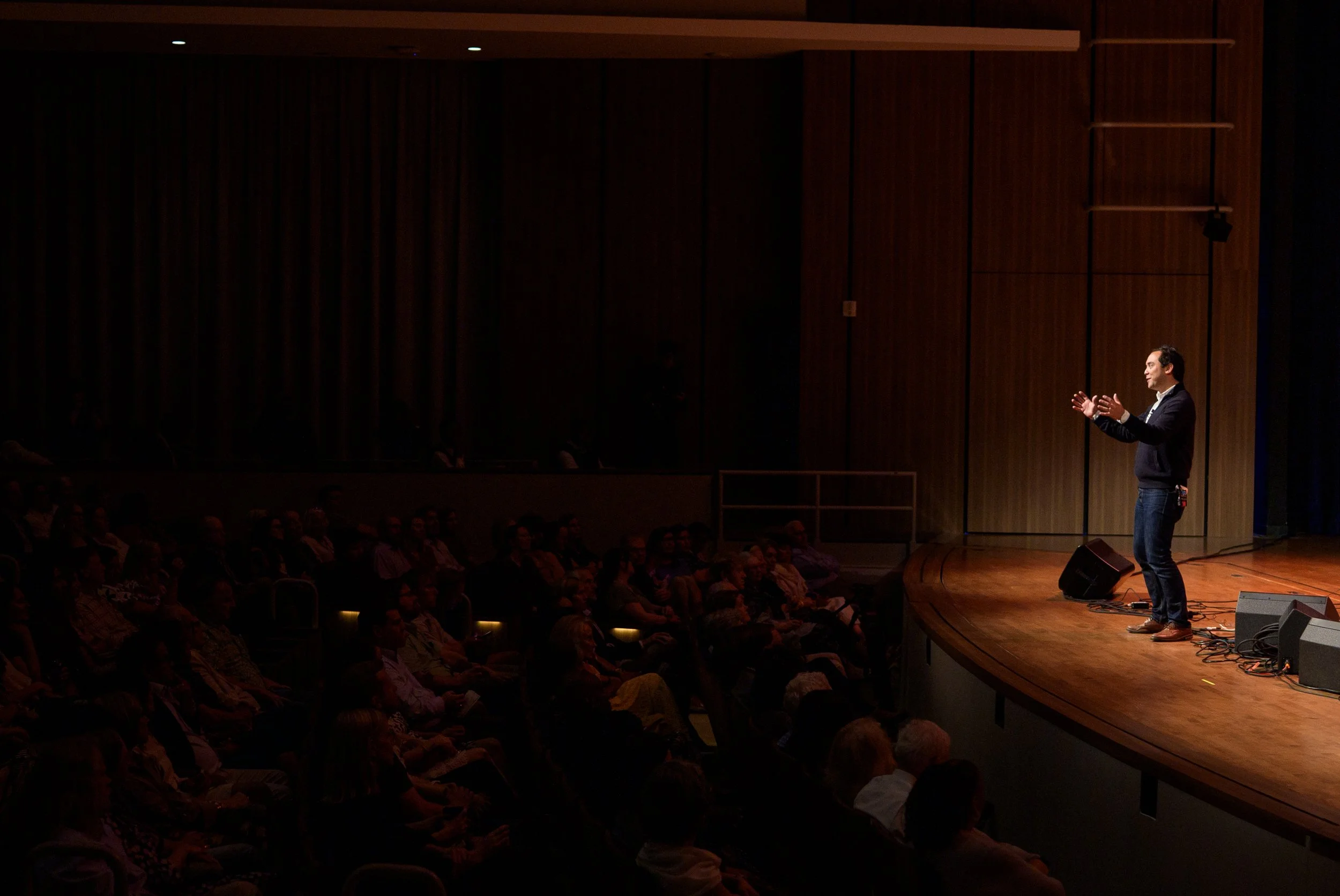 A man giving a presentation on stage in front of an audience in a darkened auditorium. The stage has a wooden floor and a brown wood-paneled wall in the background. The presenter is gesturing with his hands and appears to be engaged with the audience.