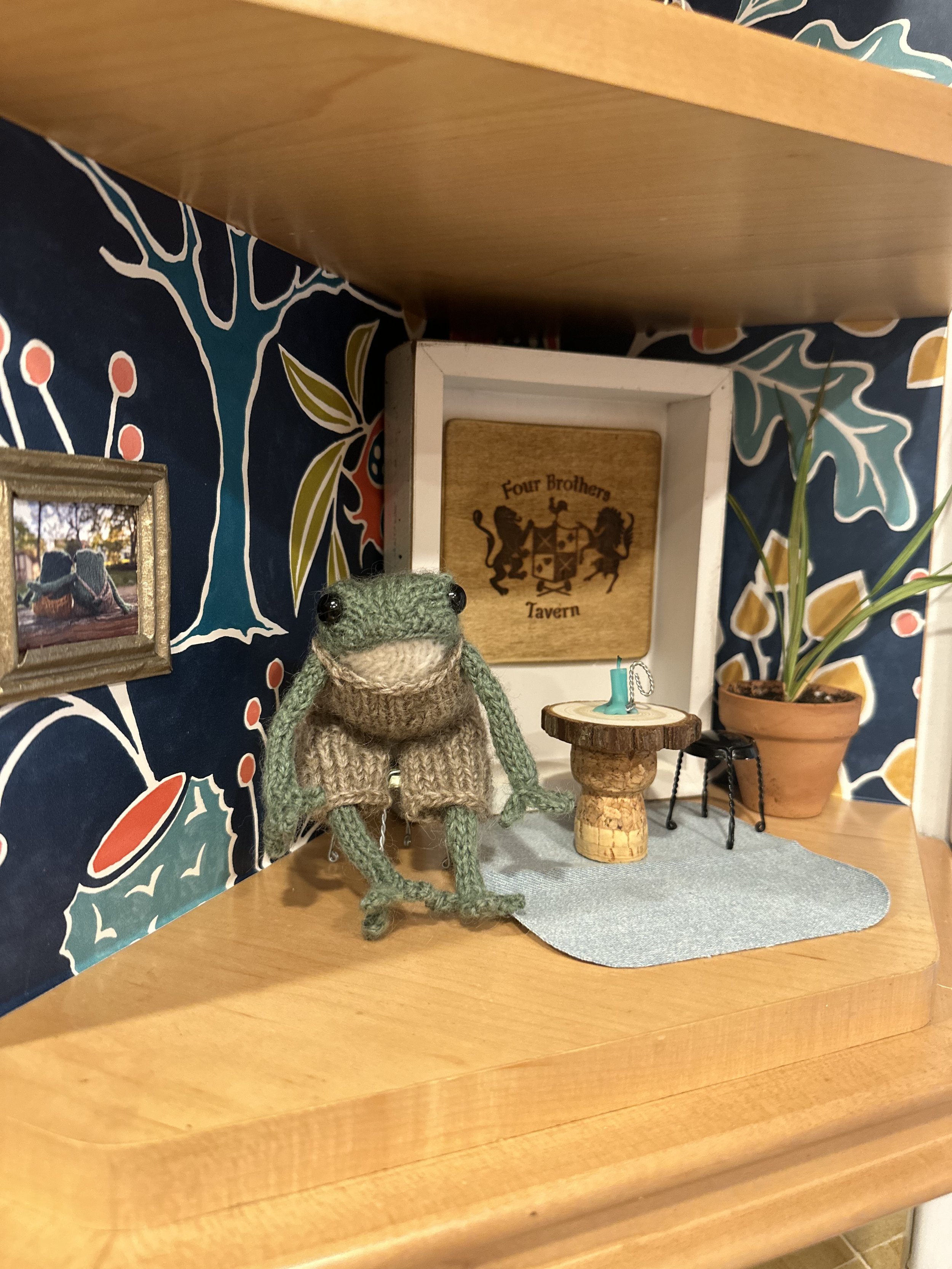 A miniature green knitted frog doll with black button eyes, positioned on a small gray mat on a wooden surface. Behind it, there is a small wooden table, a parked car photo in a golden frame, a potted plant, and a wooden sign that reads "Four Brothers Tavern" with a lion crest. The background features a colorful, abstract patterned fabric.