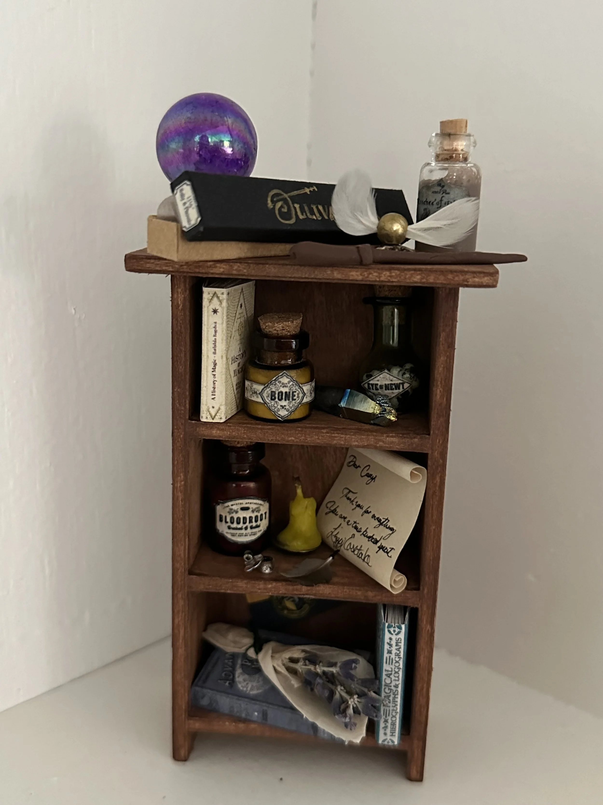 A small wooden shelf with various items on top and inside. Top shelf has a purple holographic sphere, a black box labeled 'Juju', a clear small bottle with a cork, a white feather, and a gold sphere. The middle shelf contains a book, a small jar labeled 'BONE', a small bottle labeled 'EYE & NEWT', and a scroll of paper with handwritten text. The bottom shelf has a black box, a tissue paper, a pair of rings, and a box with a dragon illustration.