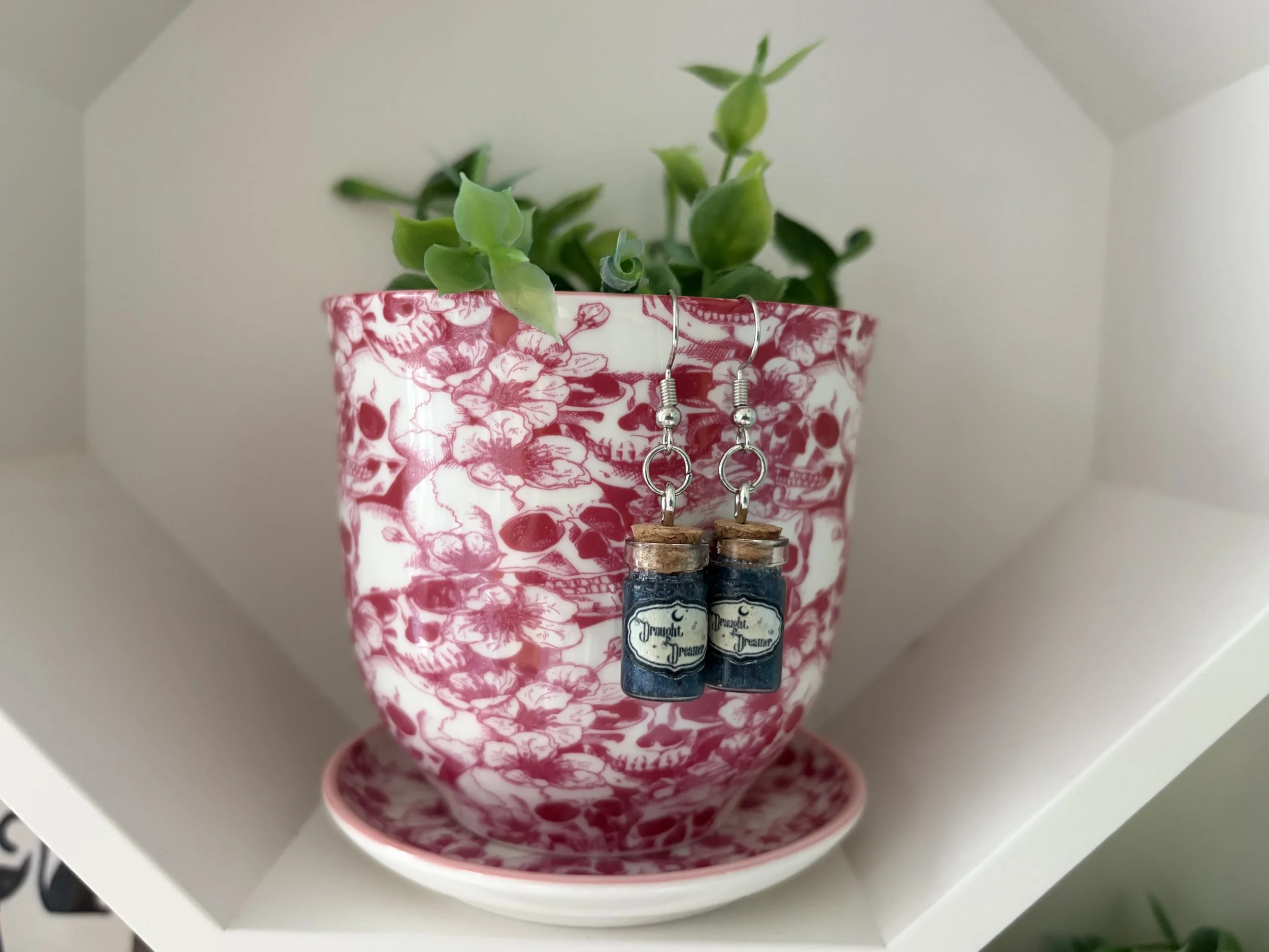 A pink mug with a skull design contains a small green leafy plant. Two small ink bottles with cork stoppers and vintage label-style designs are hanging from the mug's rim. The scene is viewed through an octagonal white frame.