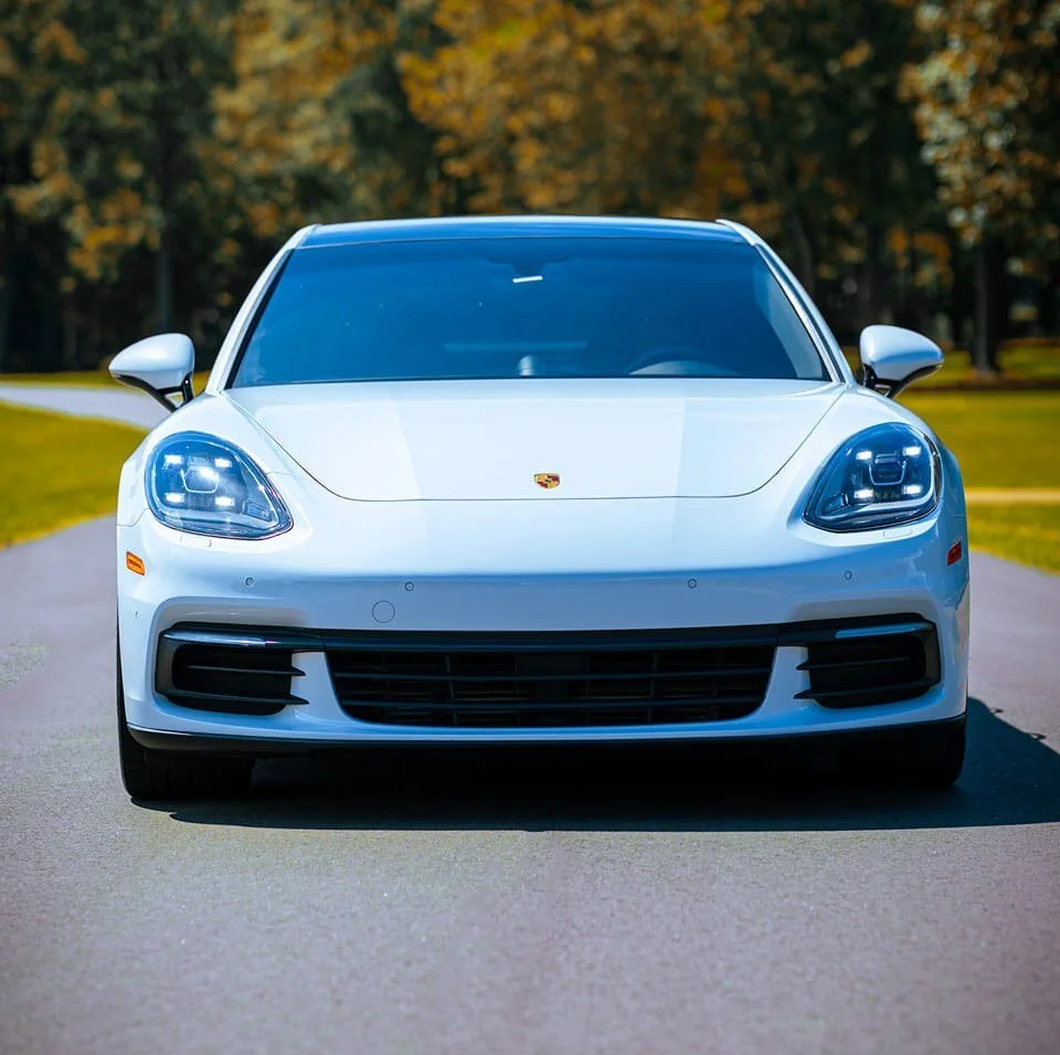 Front view of a white Porsche sports car on a paved road with trees in the background.