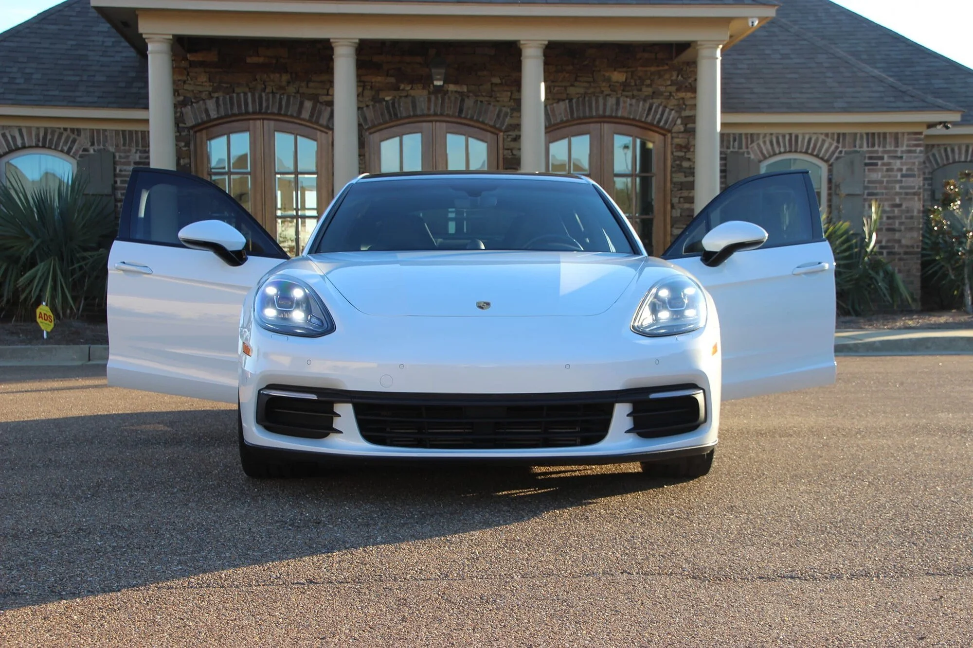 A white Porsche car with its front doors open parked in front of a brick house with large windows and columns.